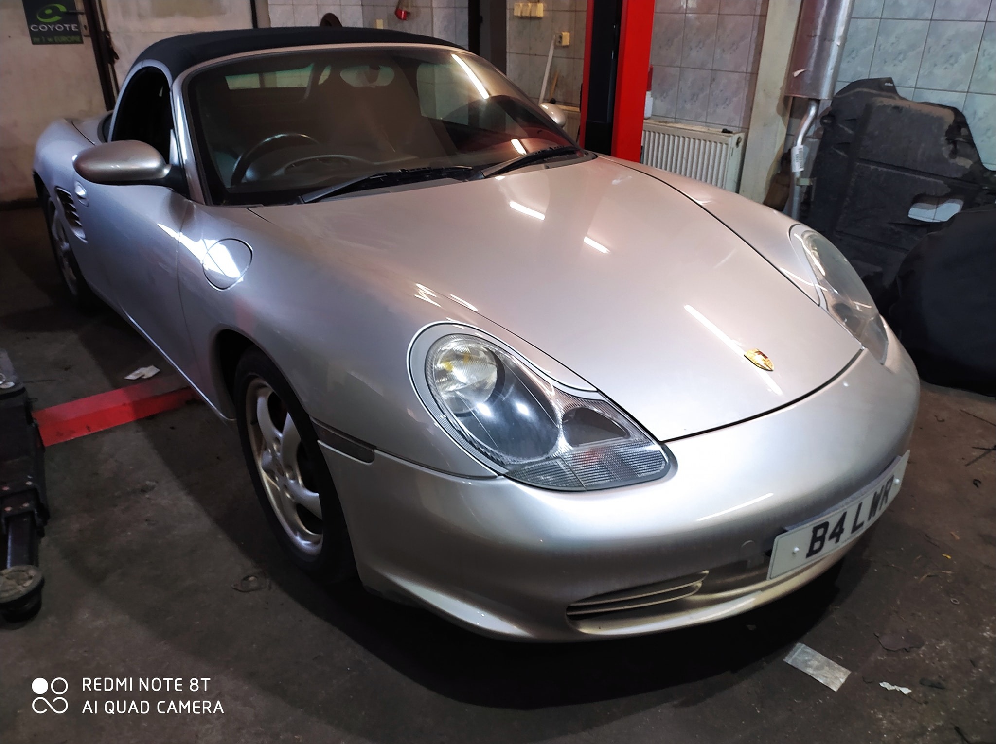 Srebrny Porsche Boxster z czarnym dachem w warsztacie, widok z przodu, tablica rejestracyjna B4 LWR, oświetlenie warsztatowe.