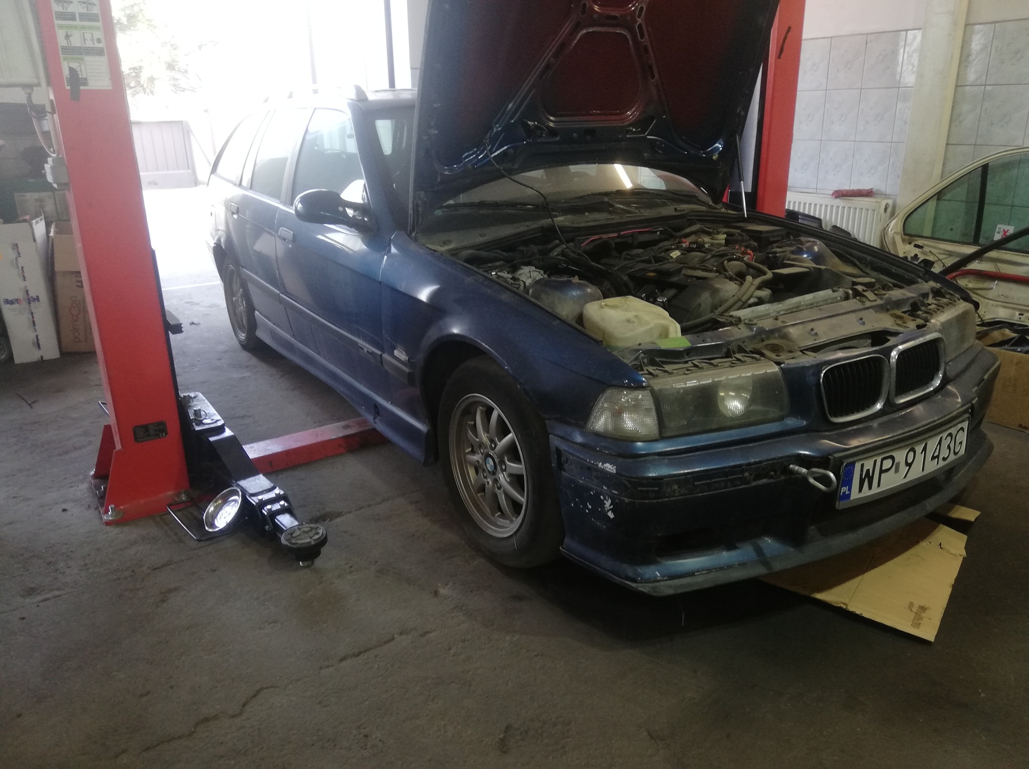 Niebieskie kombi BMW E36 na podnośniku warsztatowym z otwartą maską, widoczny silnik. Tablica rejestracyjna WP 9143G. W tle ściana warsztatu i częściowo widoczny inny pojazd.