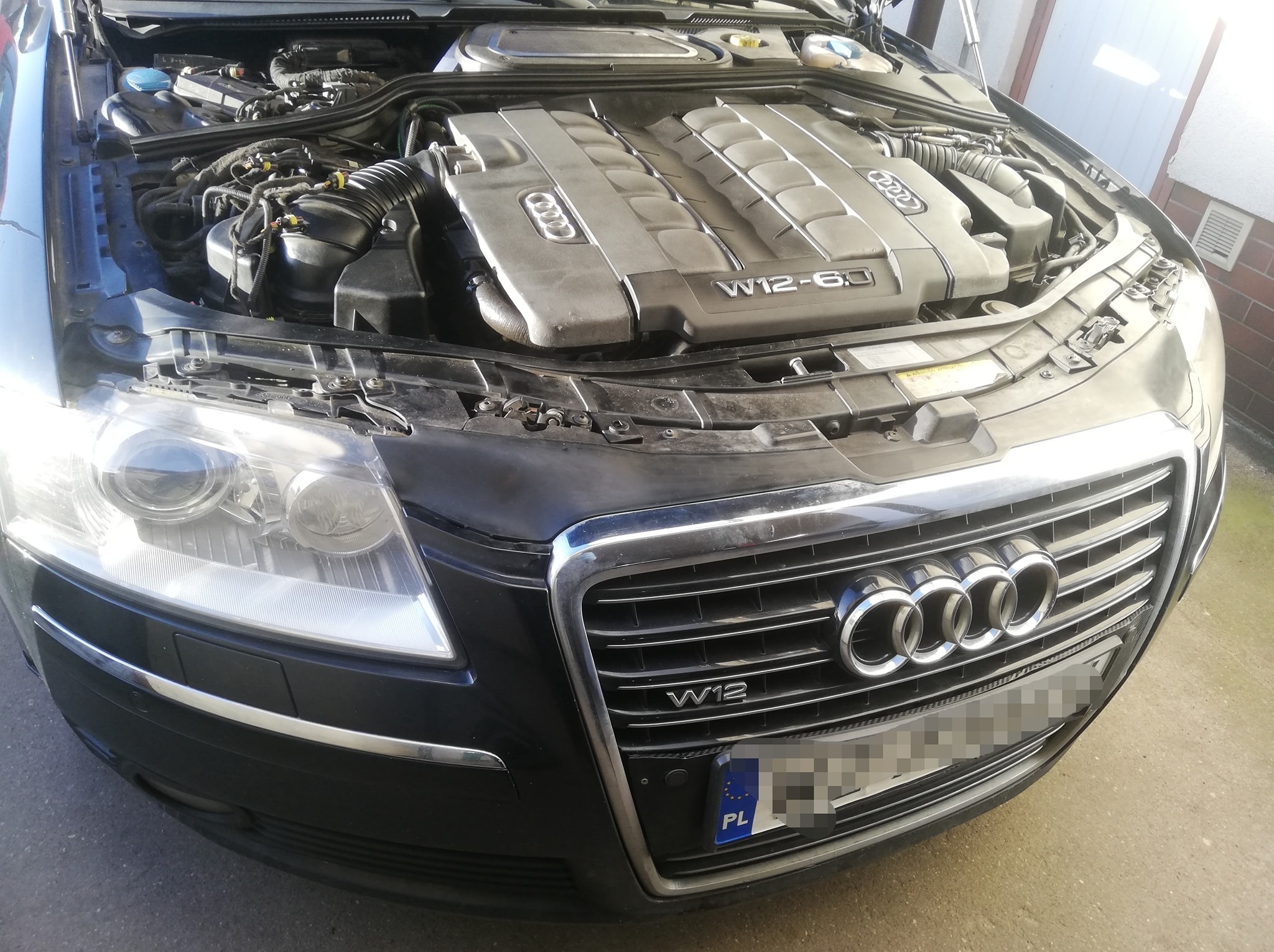 Czarny samochód Audi z otwartą maską, widoczny silnik W12 6.0. Tablica rejestracyjna zasłonięta, na grillu logo Audi i oznaczenie W12.