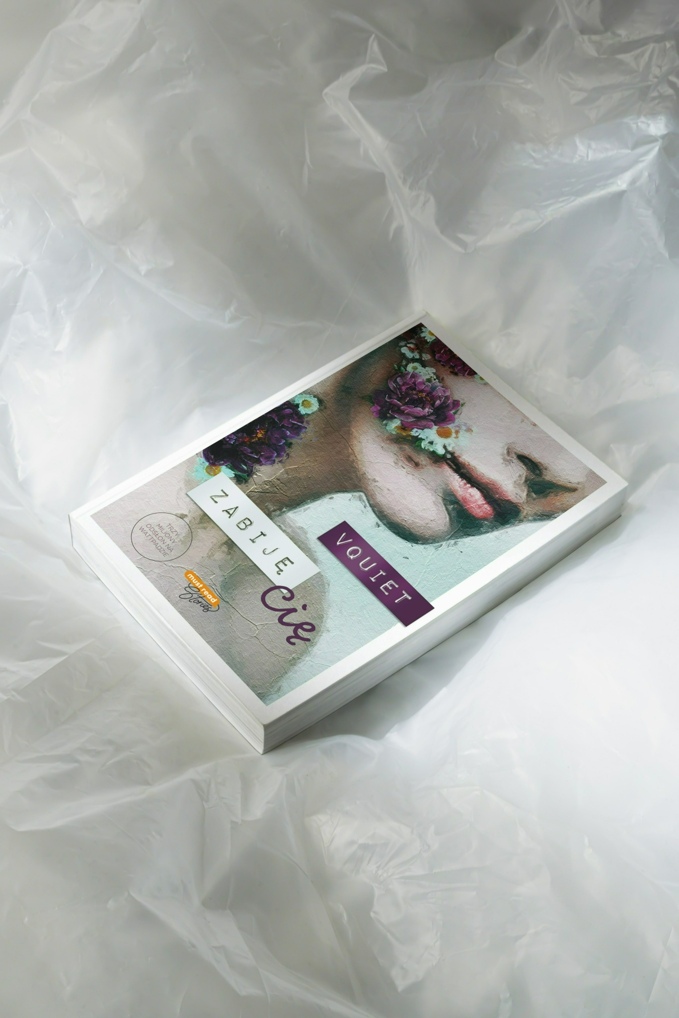 Stos okładek książek z grafiką przedstawiającą twarz z kwiatami, leżący na pogniecionej, białej folii. Ujęcie z góry, oświetlenie studyjne.