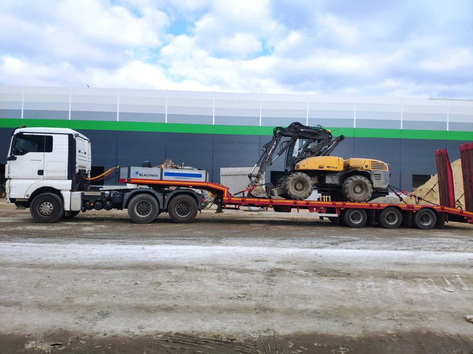 Ciężarówka laweta z żółtą koparką Mecalac na czerwonej platformie transportowej. Na naczepie widoczny osprzęt Blachmix, tło stanowi magazyn.