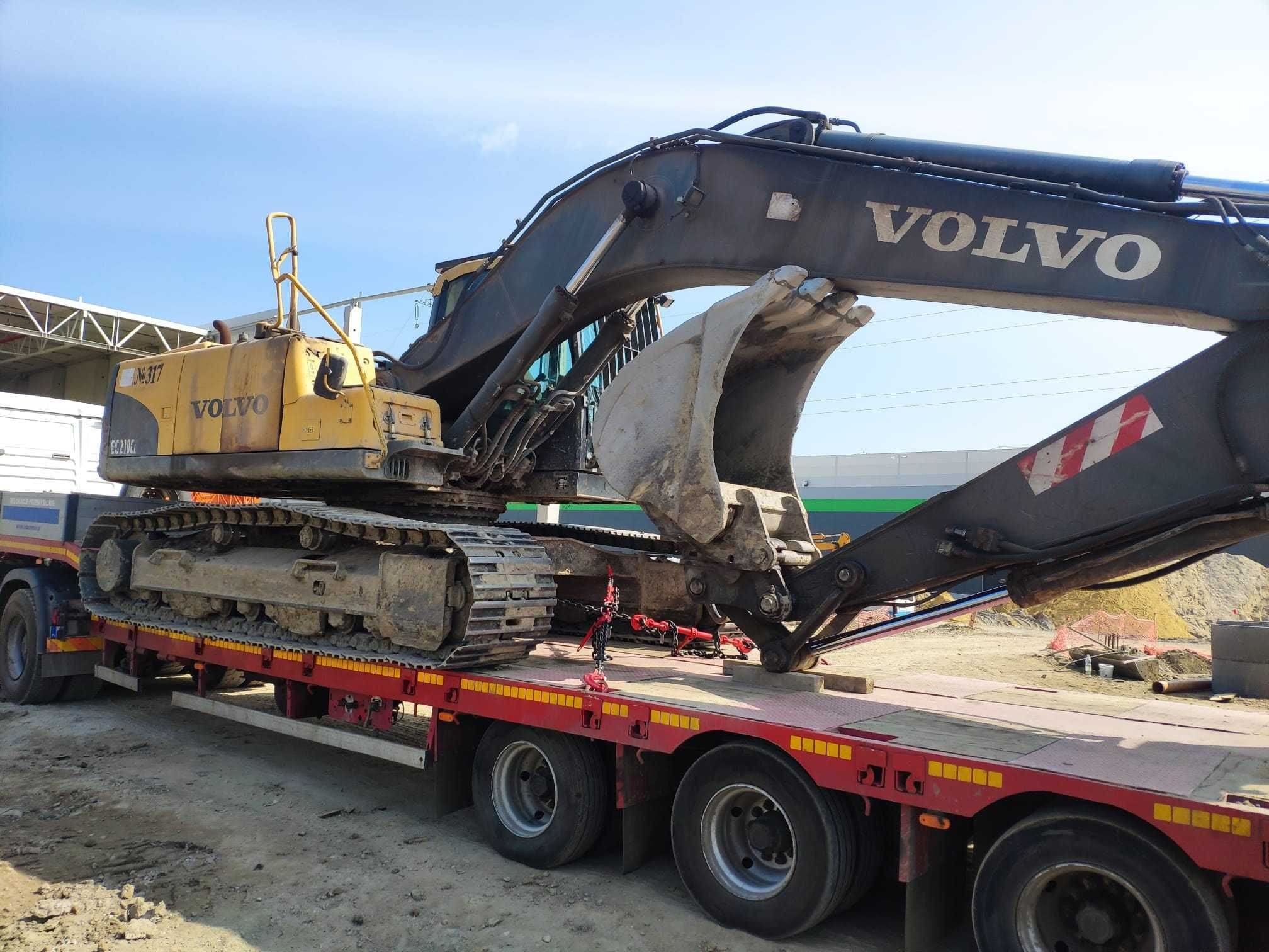 Ciężka koparka Volvo EC210CL, zabezpieczona łańcuchami, przewożona na czerwonej niskopodwoziowej lawecie. Widok z boku, w tle niebieskie niebo i hala.