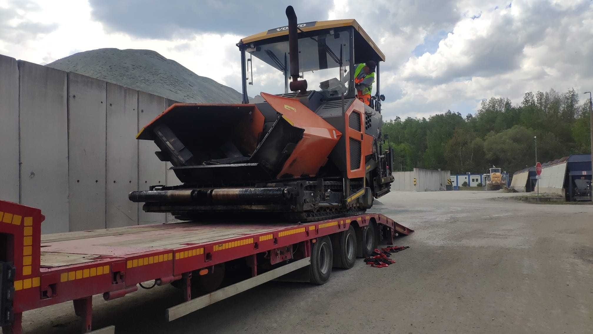 Laweta z pomarańczowym rozściełaczem asfaltu Volvo, zabezpieczonym czerwonymi pasami, na tle betonowego muru i zalesionego wzgórza.