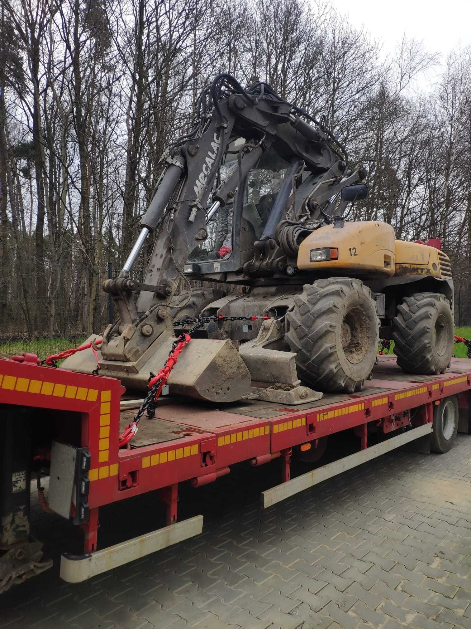 Laweta przewozi żółtą koparkę Mecalac, zabezpieczoną łańcuchami i pasami. Tło stanowią drzewa bez liści i szara kostka brukowa.