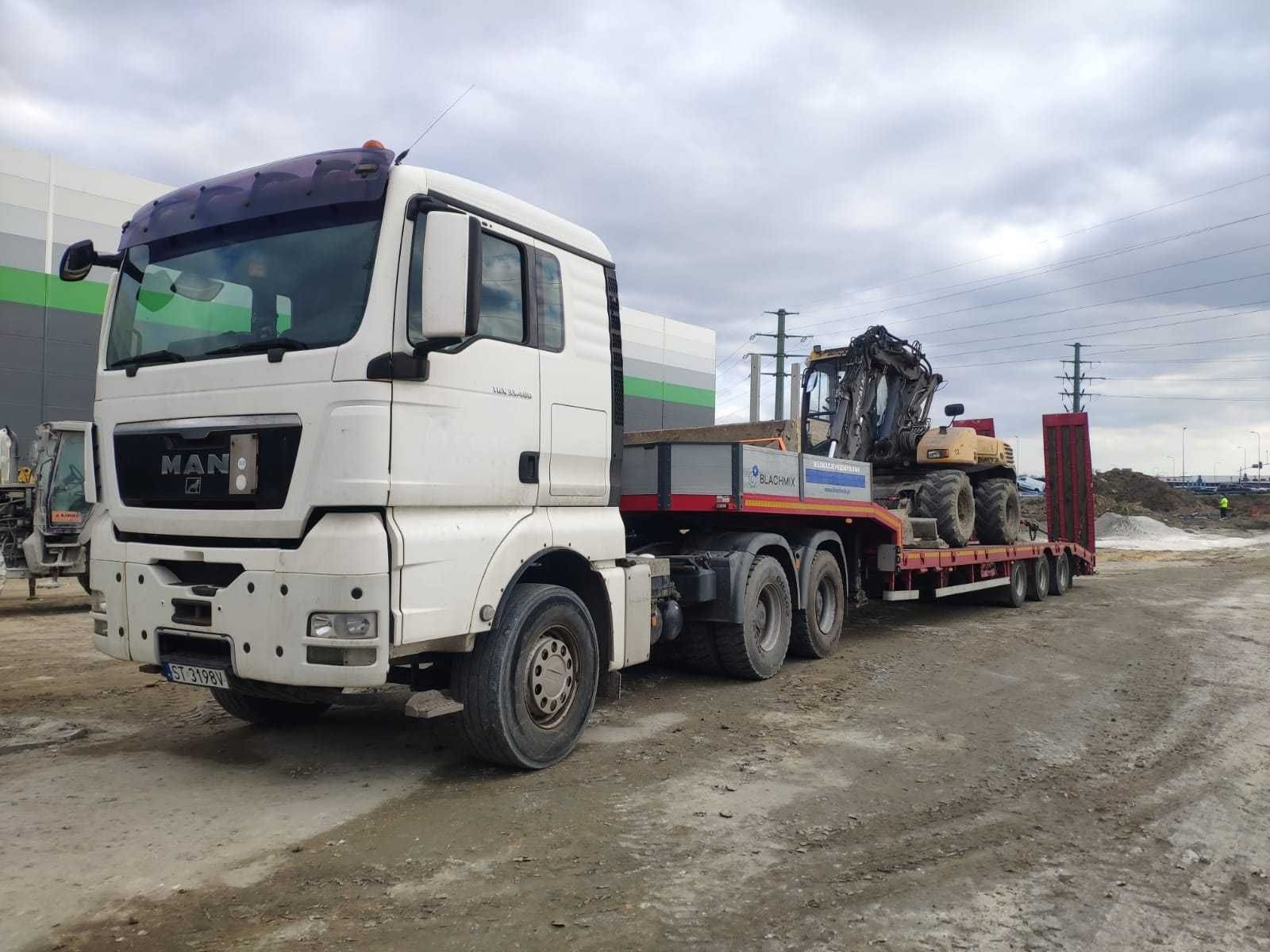 Laweta MAN z żółtą koparką na platformie, widok z przodu na placu budowy, pochmurne niebo. Tablica rejestracyjna ST 3198V.