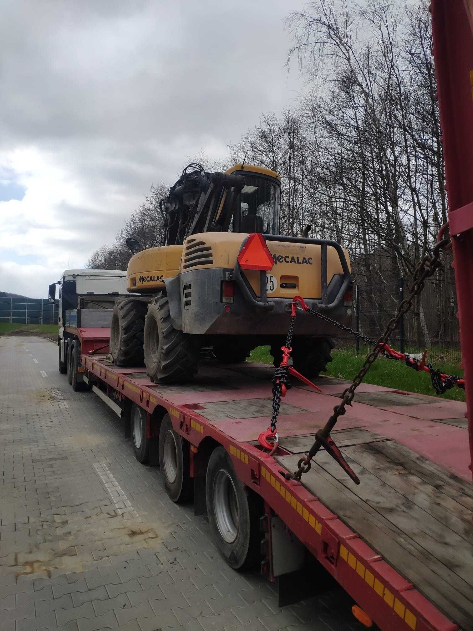 Żółta koparka Mecalac 25 na czerwonej lawecie, zabezpieczona łańcuchami, transportowana po betonowej drodze w pochmurny dzień. W tle drzewa.