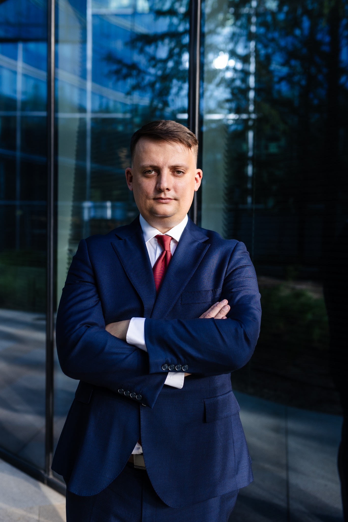 Portret mężczyzny w granatowym garniturze i czerwonym krawacie, z założonymi rękami na tle nowoczesnej szklanej fasady budynku.