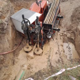 Wiert-Instal - Pomarańczowa wiertnica w wykopie wypełnionym błotem, widoczne rury wiertnicze i wystający fragment rury z zielonym oznaczeniem. Prace ziemne w toku.