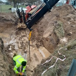 Wiert-Instal - Pracownik w jaskrawej kamizelce łączy rury w wykopie, wspierany przez maszynę do przewiertów. Widoczne błoto i elementy krajobrazu.