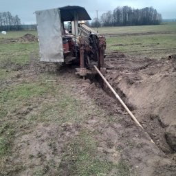 Wiert-Instal - Maszyna do robót ziemnych w trakcie wiercenia poziomego w gruncie, widoczny rów i wkładana rura. Prace ziemne na polu w pochmurny dzień.