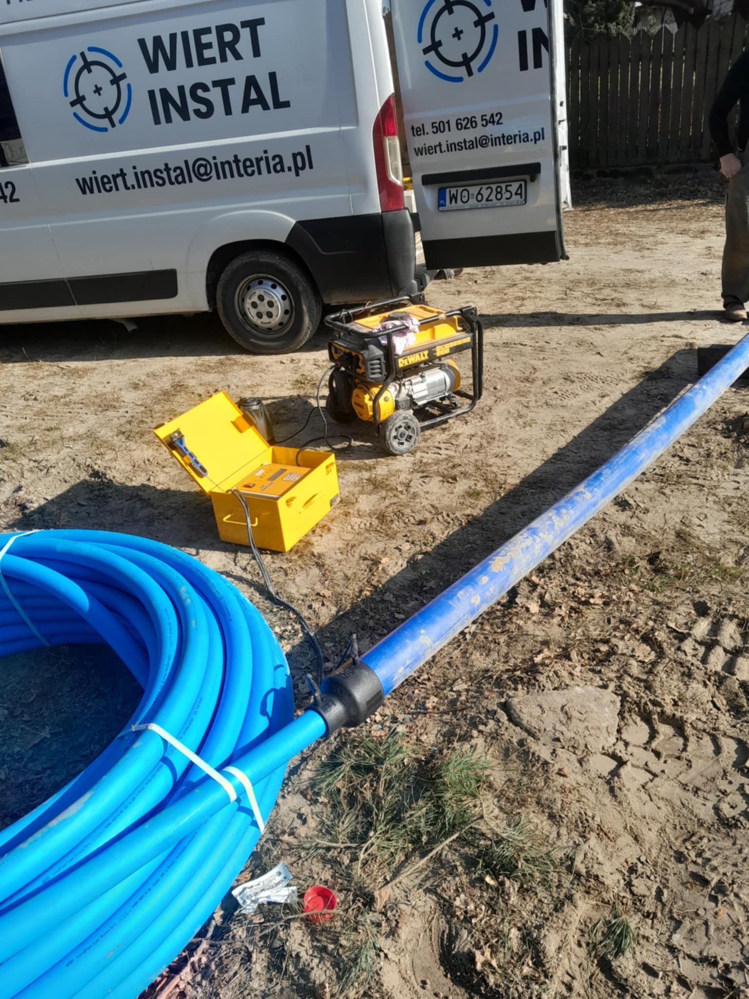 Niebieskie rury, agregat prądotwórczy DeWalt i żółta skrzynka narzędziowa na ziemi, w tle biały bus z logo firmy Wiert Instal.