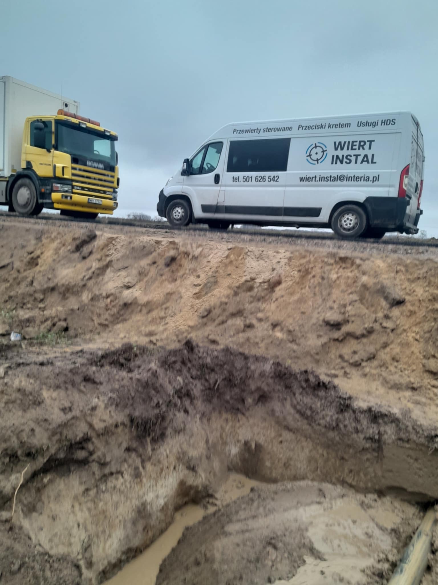 Żółta ciężarówka i biały bus firmy Wiert Instal na tle wykopu ziemnego. Przewierty sterowane, przeciski kretem i usługi HDS.