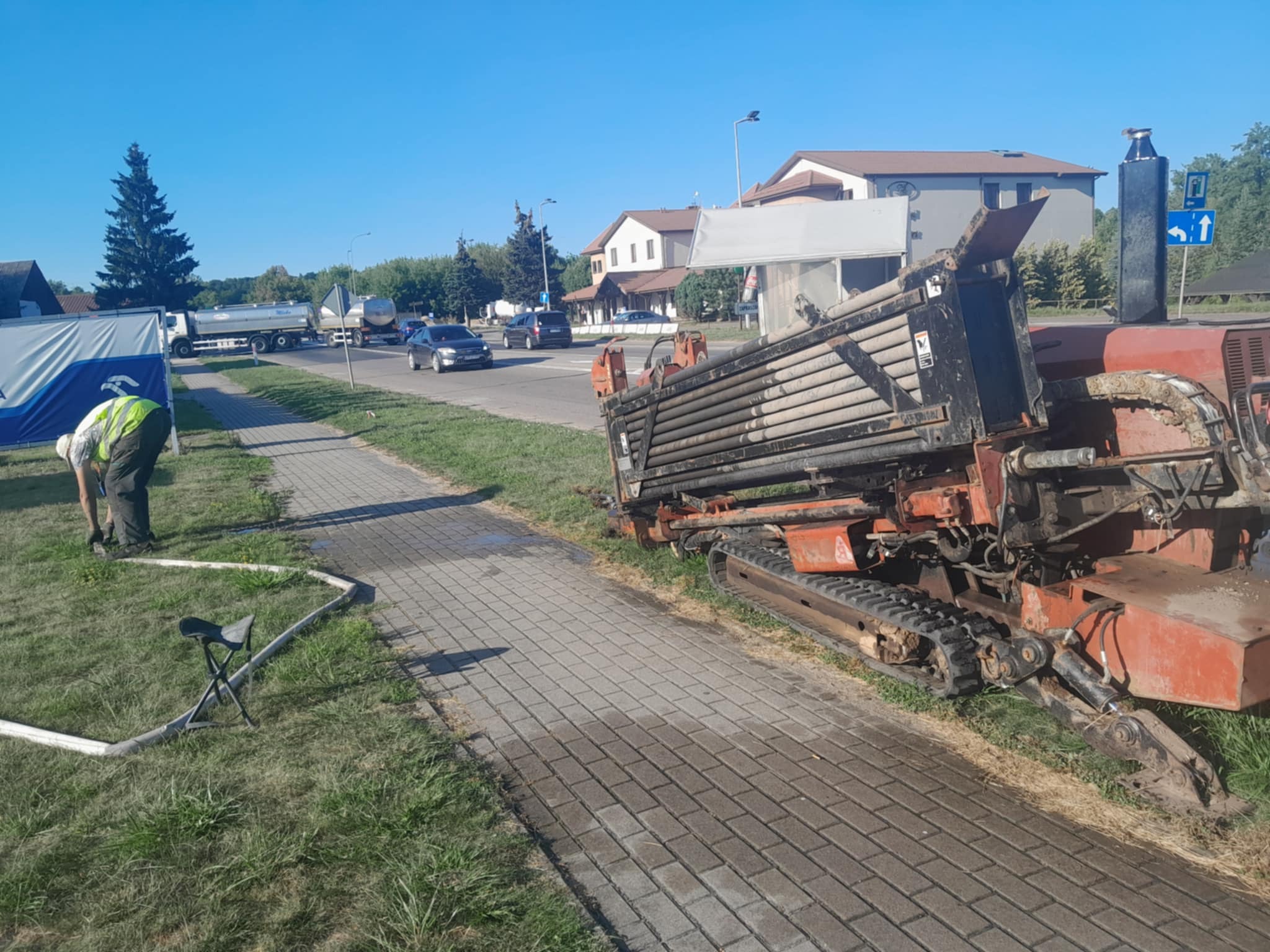 Pomarańczowa wiertnica gąsienicowa na chodniku z kostki, pracownik w odblaskowej kamizelce przy białym wężu. W tle ulica z autami i zabudowania.