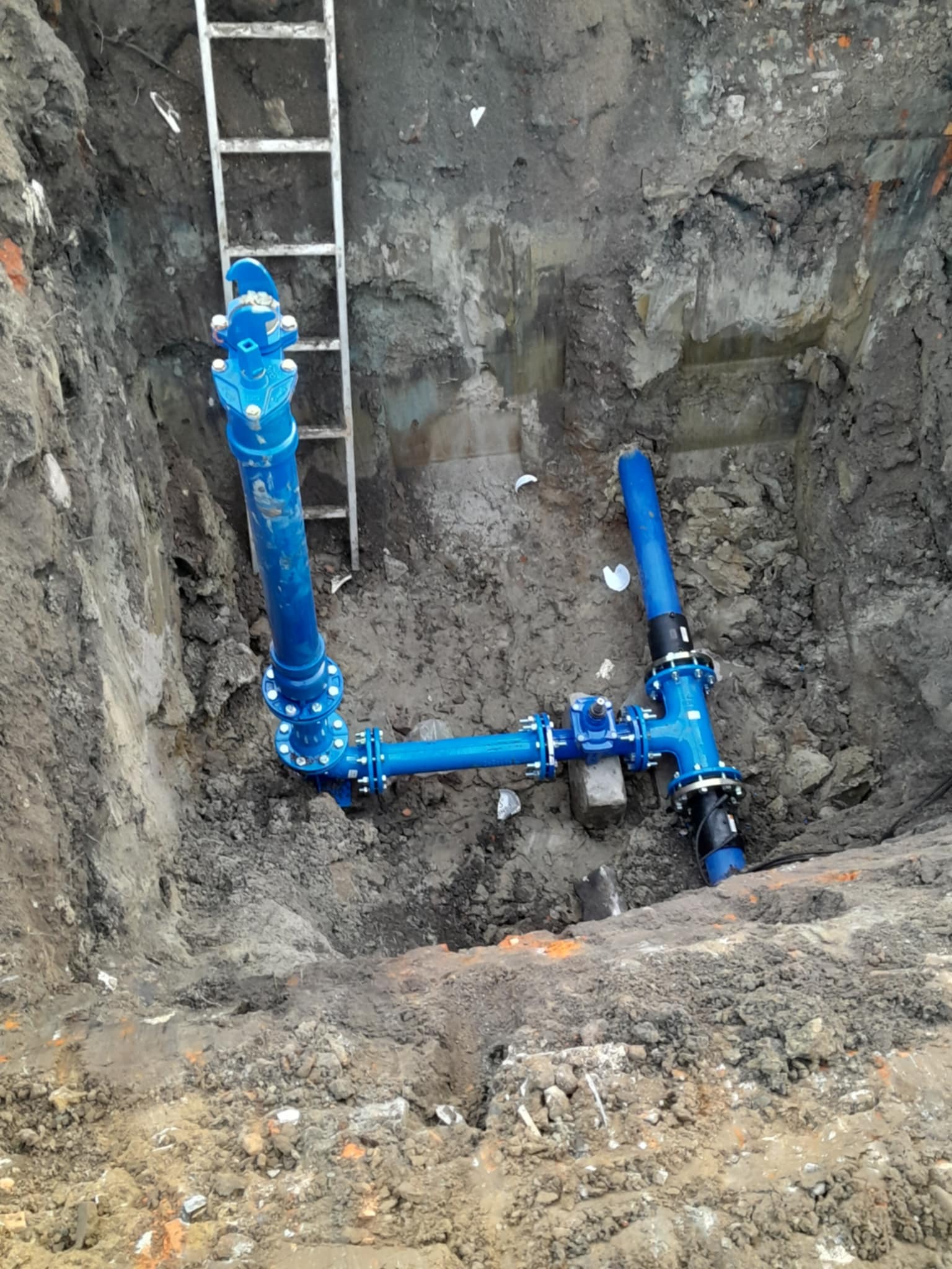 Niebieskie rury połączone w wykopie ziemnym z drabiną, prawdopodobnie instalacja wodociągowa lub kanalizacyjna.