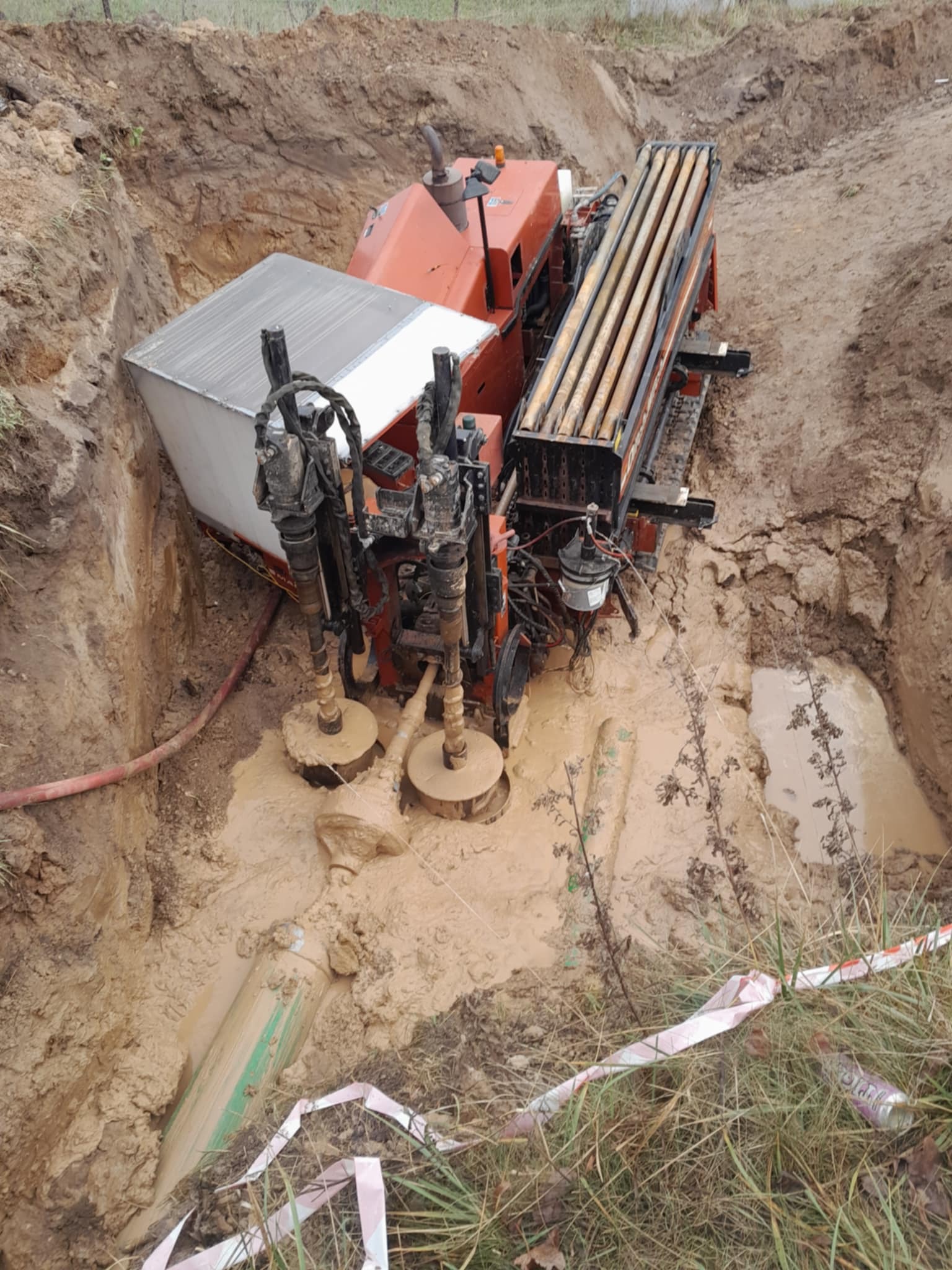 Pomarańczowa wiertnica w wykopie wypełnionym błotem, widoczne rury wiertnicze i wystający fragment rury z zielonym oznaczeniem. Prace ziemne w toku.