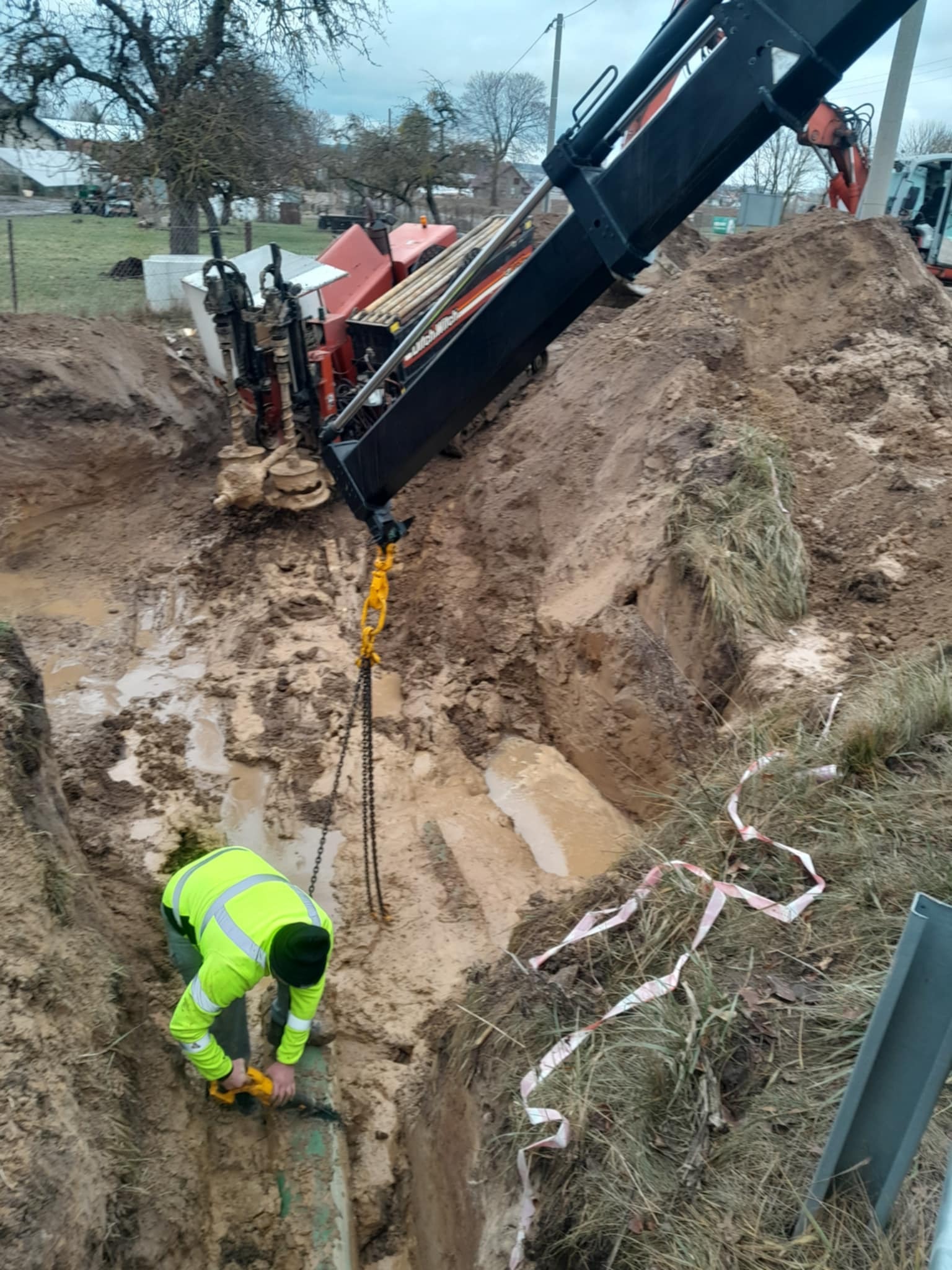 Pracownik w jaskrawej kamizelce łączy rury w wykopie, wspierany przez maszynę do przewiertów. Widoczne błoto i elementy krajobrazu.