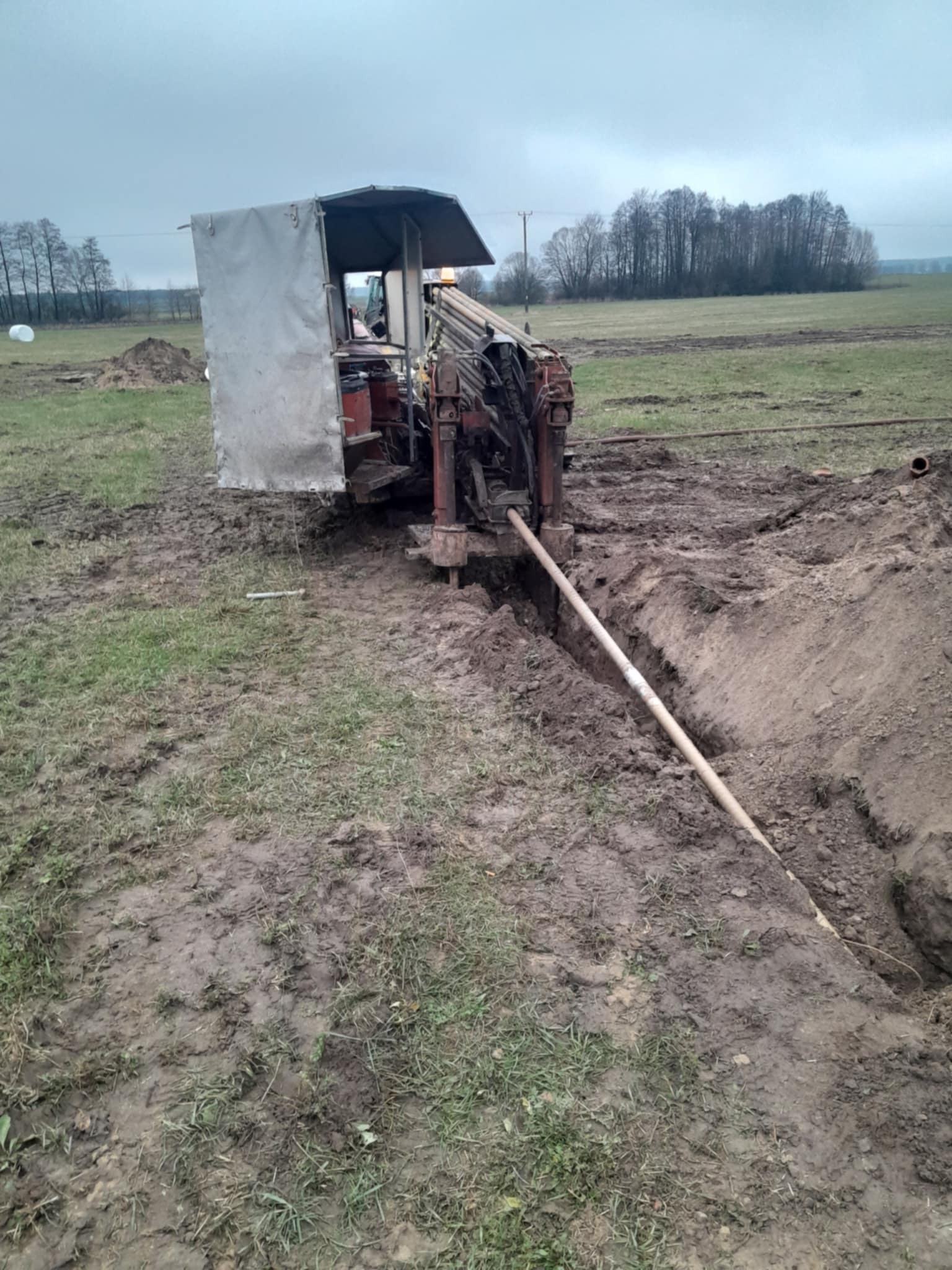 Maszyna do robót ziemnych w trakcie wiercenia poziomego w gruncie, widoczny rów i wkładana rura. Prace ziemne na polu w pochmurny dzień.