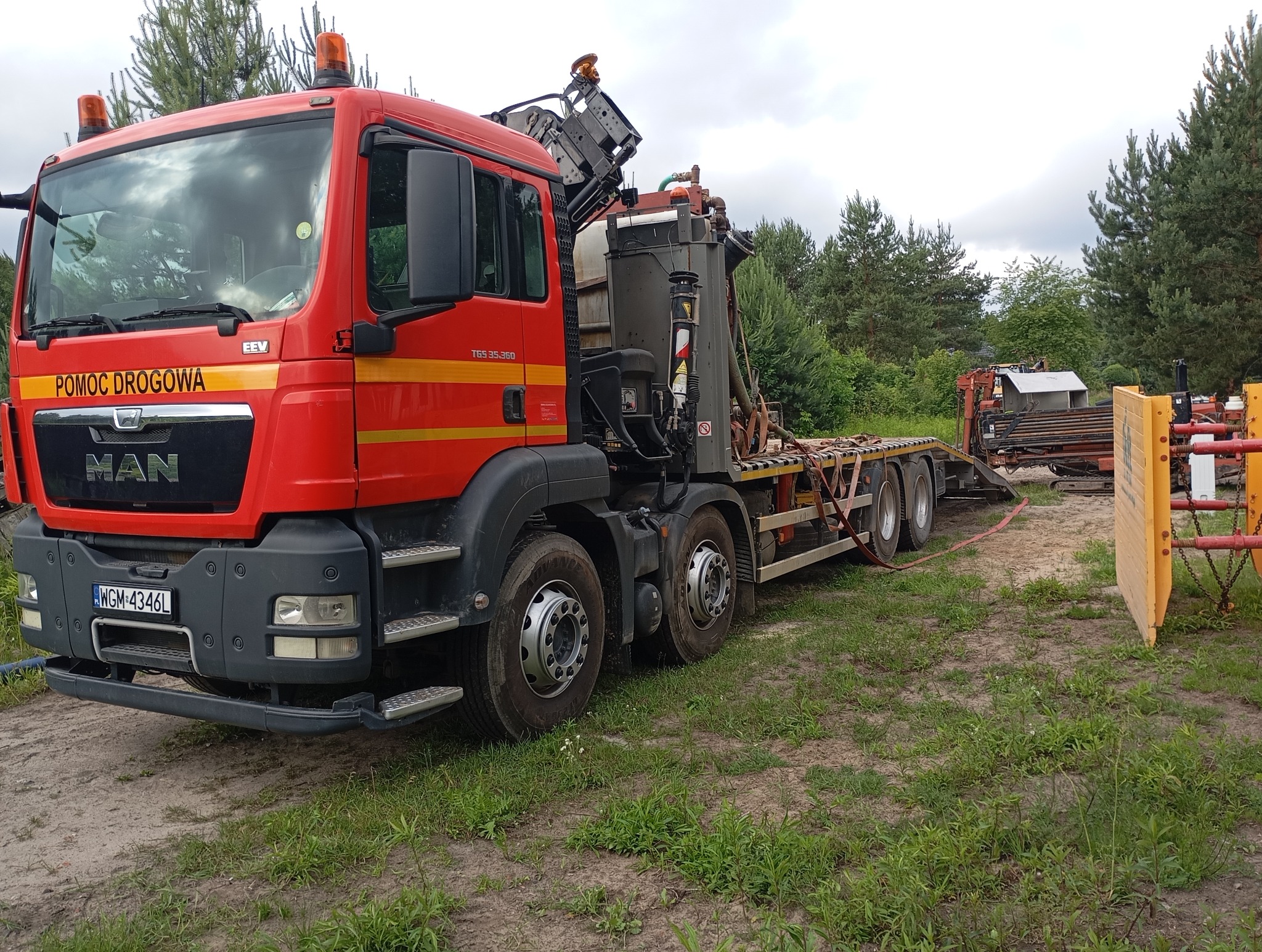 Czerwony samochód ciężarowy MAN z napisem 'Pomoc Drogowa' i specjalistycznym sprzętem na naczepie, prawdopodobnie do przewiertów, stoi na trawiastym terenie.