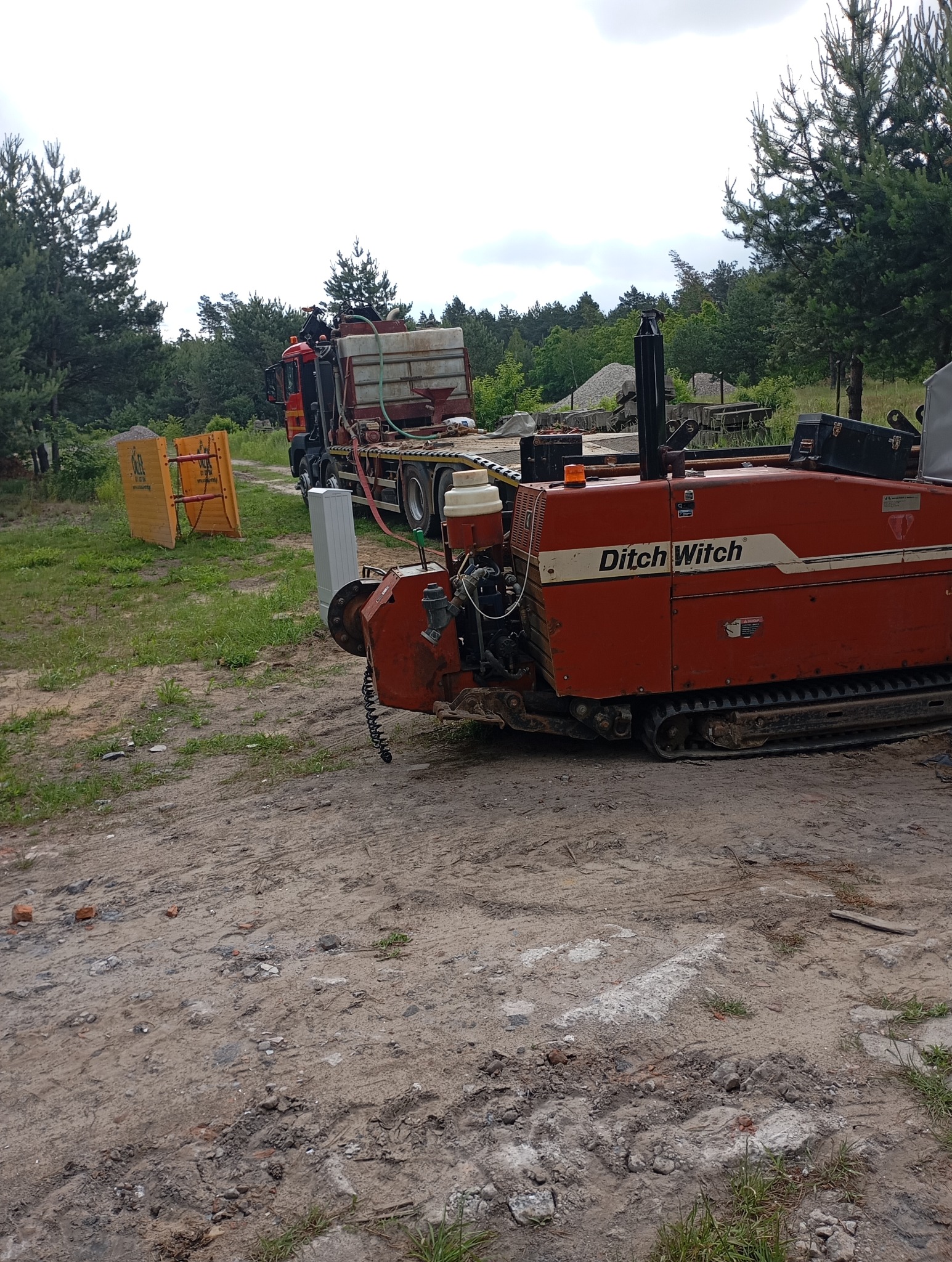 Pomarańczowa maszyna Ditch Witch na gąsienicach, na ziemi, z ciężarówką w tle. Sprzęt do przewiertów na placu budowy.