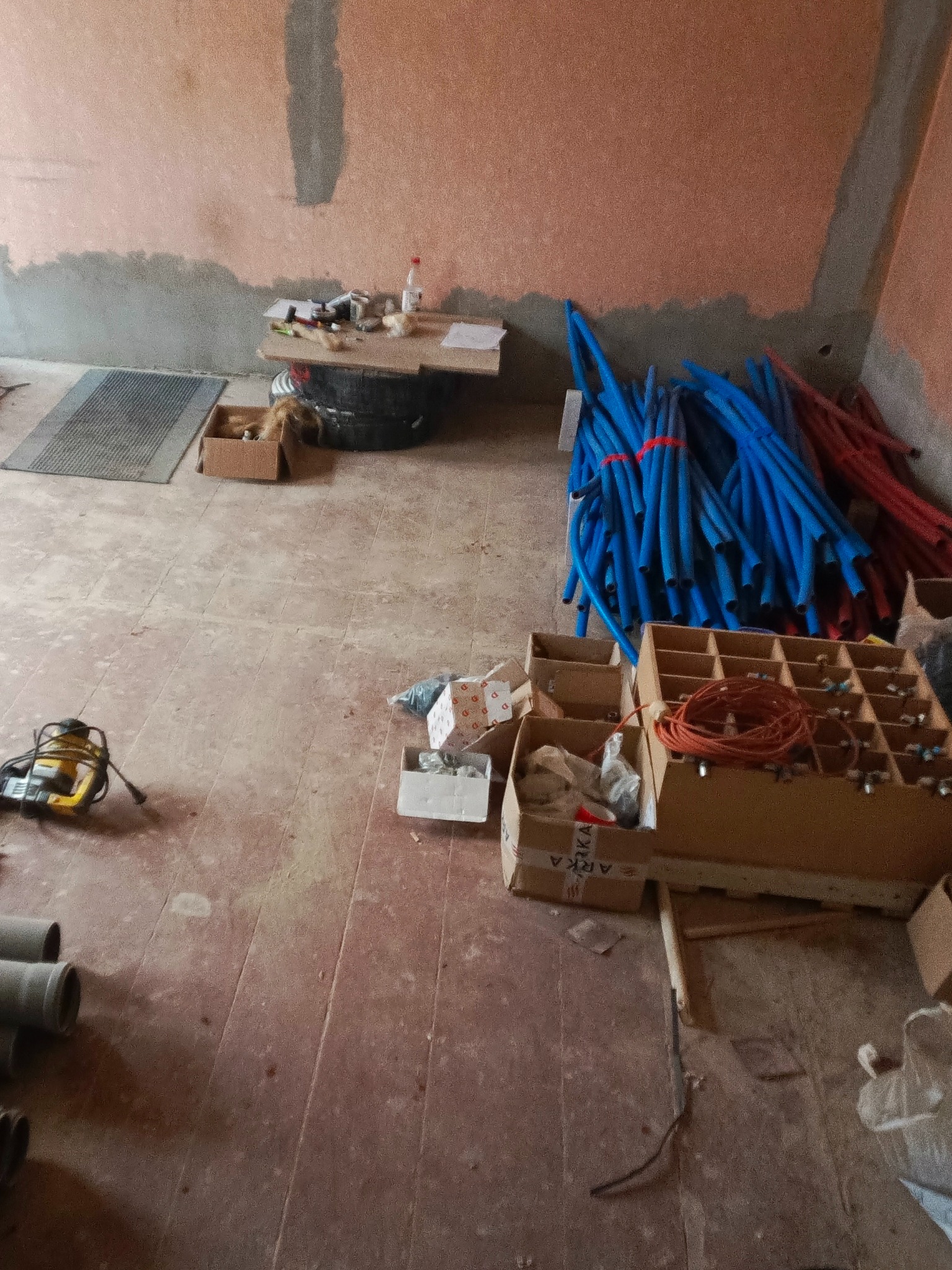 Przygotowania do instalacji: niebieskie i czerwone rury, narzędzia, kartony z akcesoriami na podłodze w surowym wnętrzu przed wykończeniem ścian.