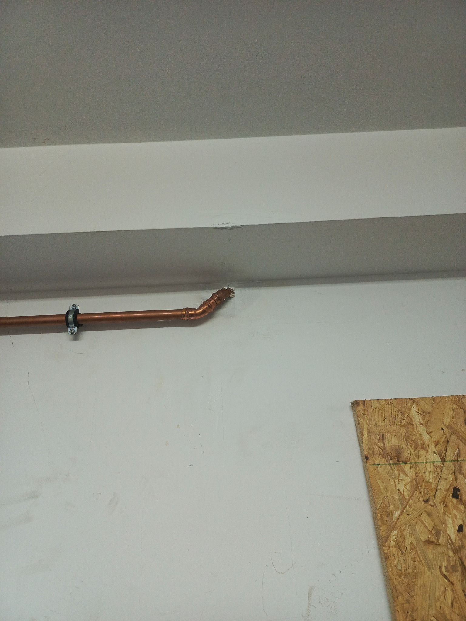 Miedziana rura instalacyjna, zamocowana do ściany klamrą, z widocznym kolankiem i lutowanymi łączeniami, na tle białej ściany i sufitu. Część płyty OSB w prawym dolnym rogu.