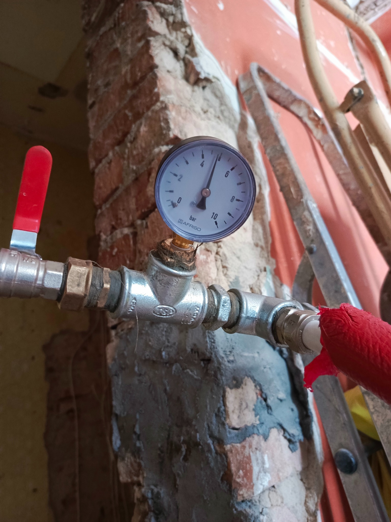 Metalowa instalacja z manometrem na tle ceglanej ściany w trakcie remontu. Czerwony zawór i fragment drabiny w tle. Widoczne ślady po kuciu w ścianie.