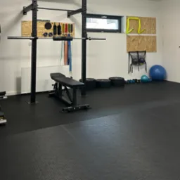 Domowa siłownia z czarną podłogą, sprzętem do ćwiczeń, takim jak atlas, ławeczka, hantle i piłki, oraz akcesoriami zawieszonymi na ścianie.