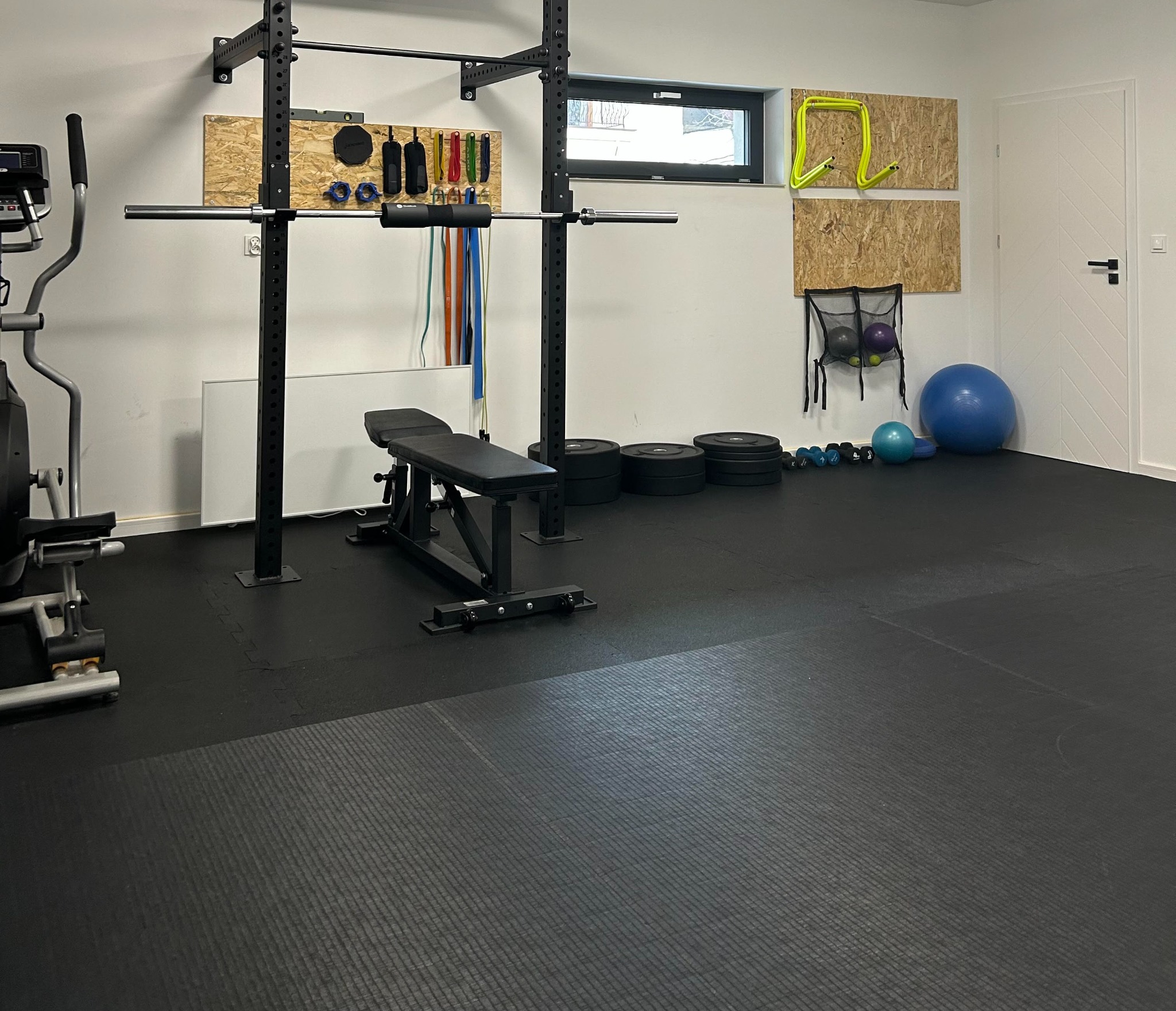 Domowa siłownia z czarną podłogą, sprzętem do ćwiczeń, takim jak atlas, ławeczka, hantle i piłki, oraz akcesoriami zawieszonymi na ścianie.