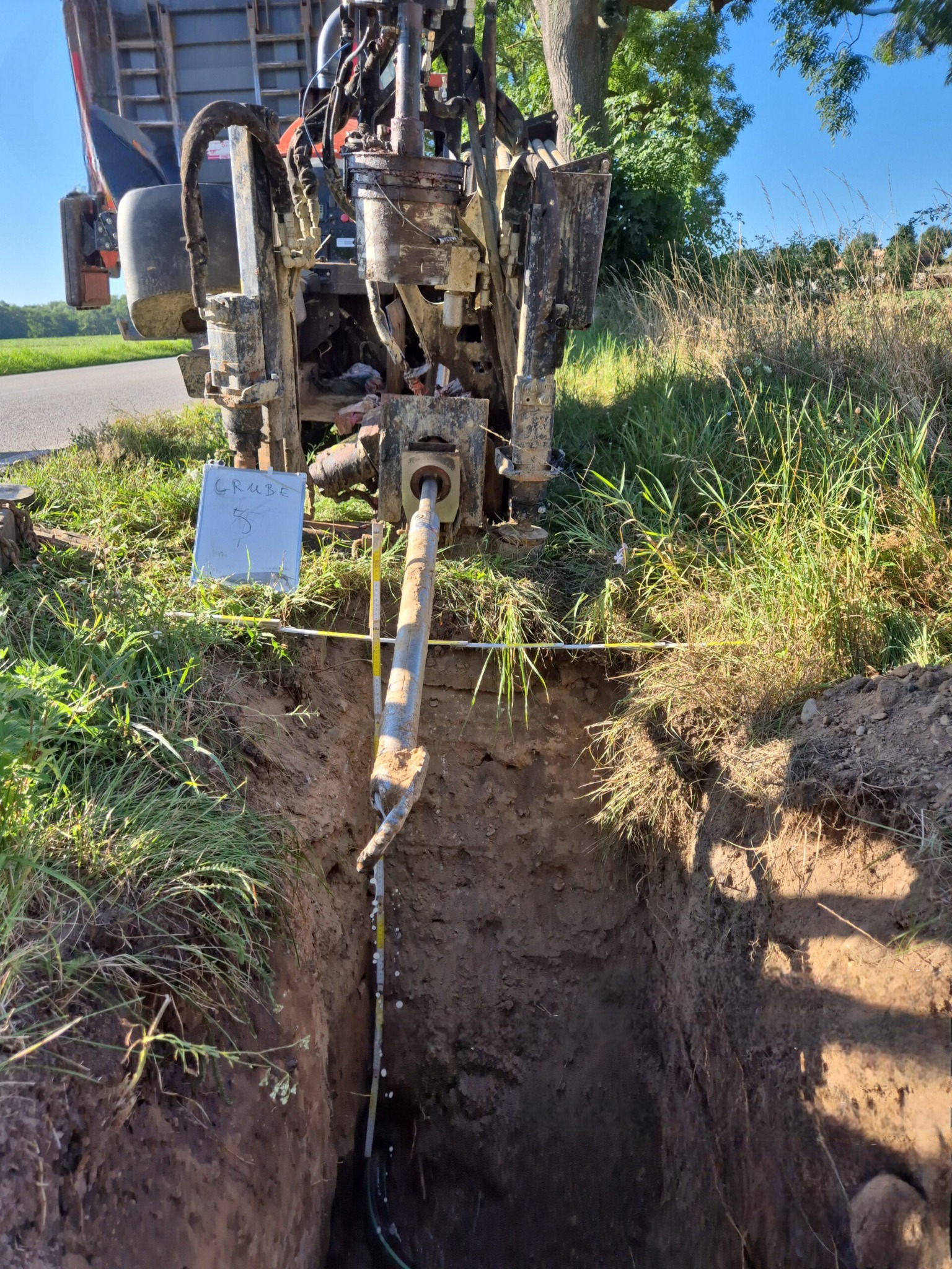 Maszyna do robót ziemnych zagłębia się w wykop, widoczna rura w ziemi. Na pierwszym planie tabliczka z napisem 'Grube 5'.