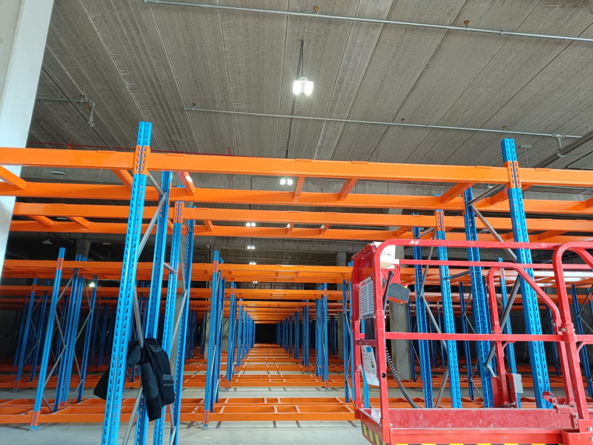 Montaż regałów magazynowych w Jabłonnie. Widok na pomarańczowo-niebieską konstrukcję z podnośnika. Perspektywa w głąb hali, oświetlenie sufitowe.