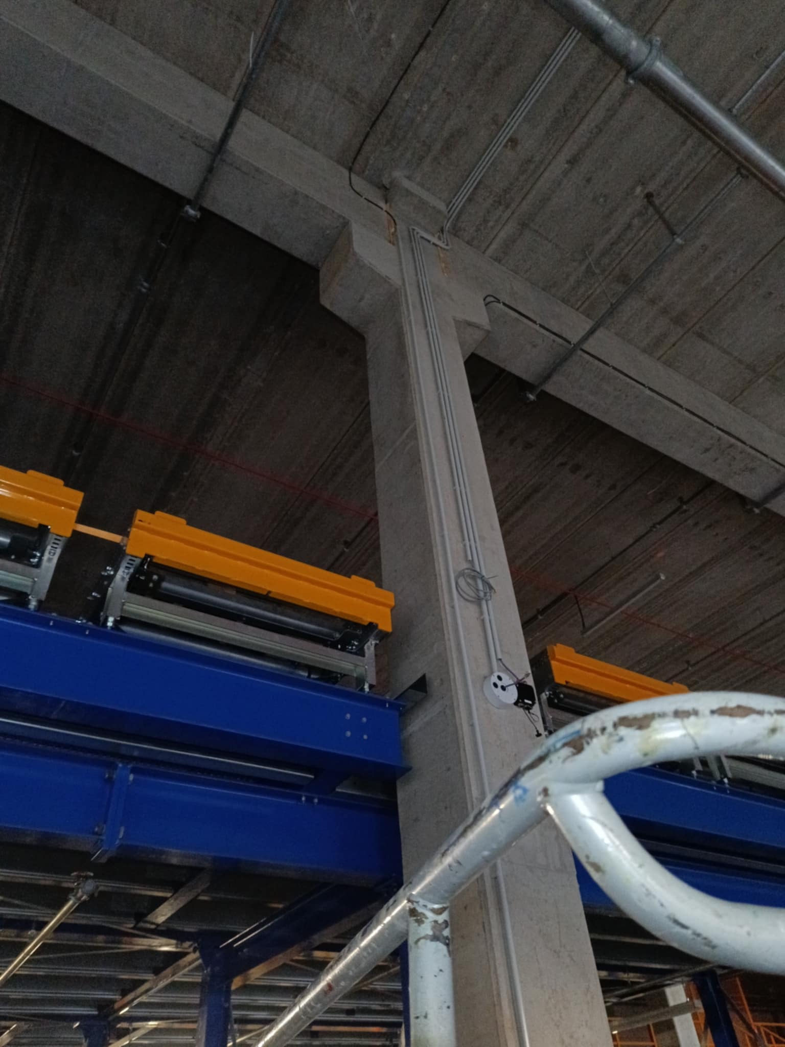 Szare, surowe wnętrze z betonowym filarem i widocznymi instalacjami elektrycznymi. Elementy konstrukcji stalowej w kolorze niebieskim i żółtym.