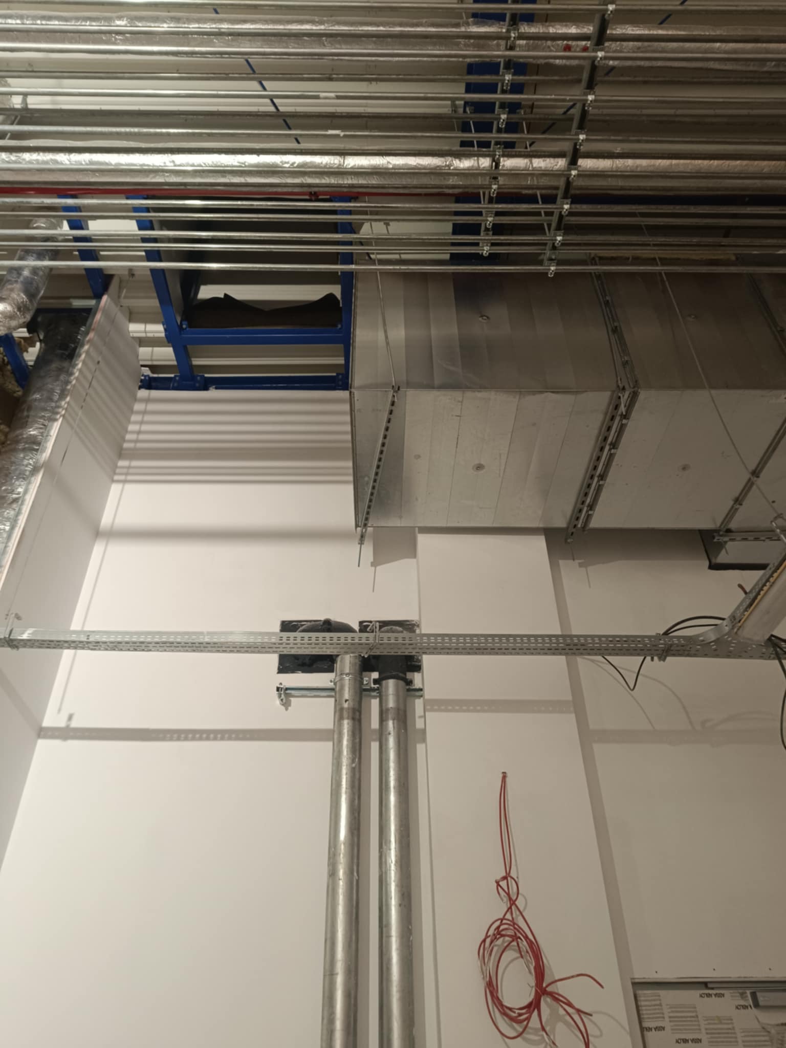 Biała ściana z instalacją wentylacyjną, rurami i kablami. Widoczne rury pod sufitem i instalacja wentylacyjna. Kabel zwisa ze ściany. Nowe tynki.