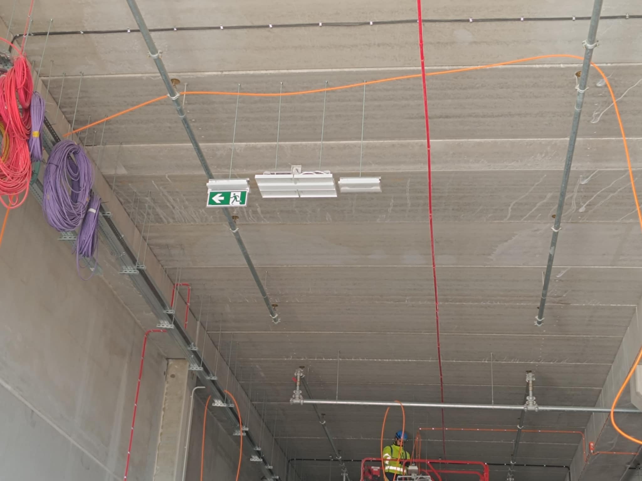 Instalacje elektryczne i rury pod sufitem z betonu. Widoczne kable w kolorach: czerwonym, pomarańczowym, fioletowym i żółtym. Zamontowane lampy i znaki ewakuacyjne.