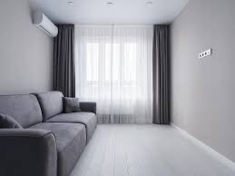 Minimalistyczny salon w odcieniach szarości: sofa, zasłony, klimatyzator. Jasna podłoga i okno z białymi firanami tworzą spokojną atmosferę.