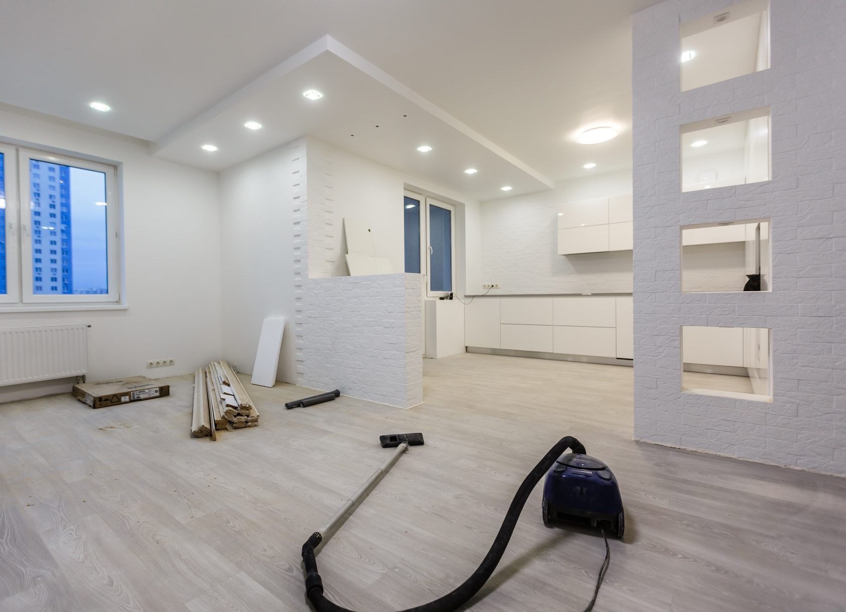 Jasne wnętrze po remoncie: parkiet, białe ściany z cegły, kuchnia w tle. Odkurzacz na pierwszym planie, resztki materiałów budowlanych przy oknie.