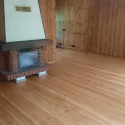 WW Parkiety - Jasne, świeżo wycyklinowane deski podłogowe w pokoju z boazerią i kominkiem. Widoczne detale: ceglana obudowa kominka, drewniany sufit z ciemnymi belkami.