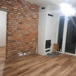 WW Parkiety - Jasne panele podłogowe w pokoju z ceglaną ścianą, białym kominkiem i dużymi oknami balkonowymi. Widoczny drewniany stołek i przejście do innego pomieszczenia.