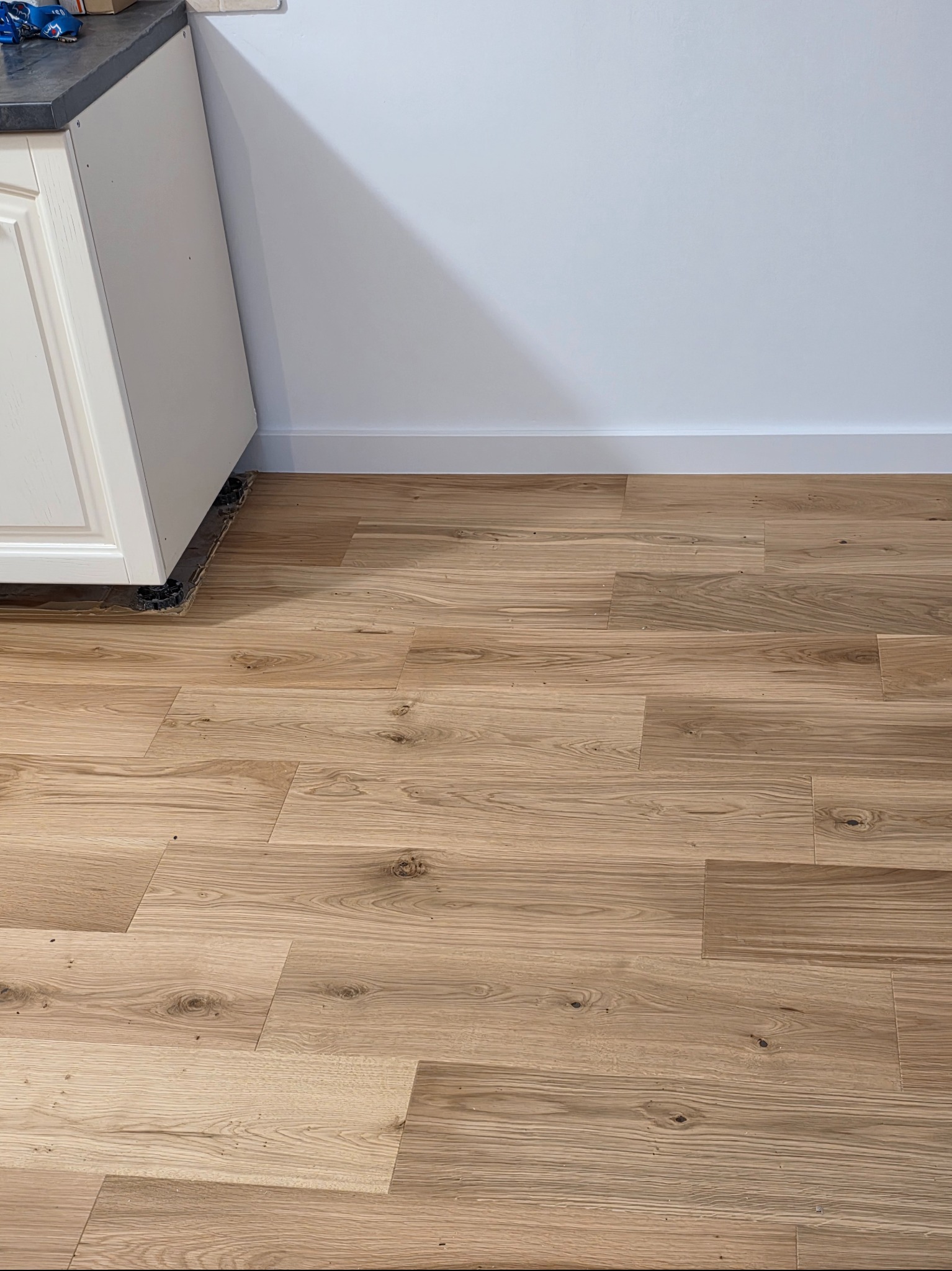 Ułożone panele podłogowe z widocznym wzorem drewna, przy białej ścianie i szafce kuchennej. Widoczne detale wykończenia podłogi przy meblach.