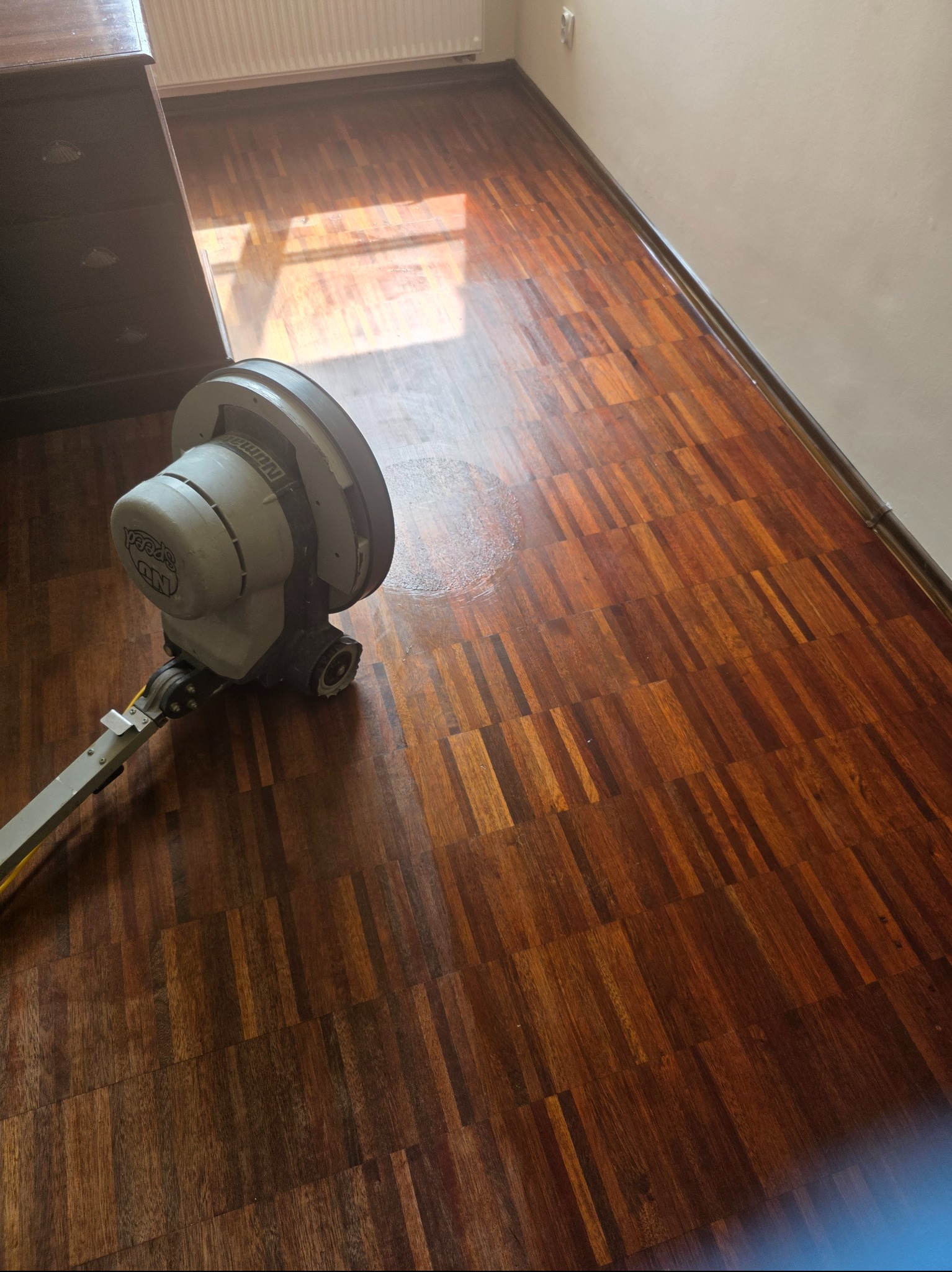 Parkiet po cyklinowaniu z widoczną maszyną do cyklinowania. Ciemne drewno z wyraźnym usłojeniem, błyszczące wykończenie. Fragment komody w lewym górnym rogu.