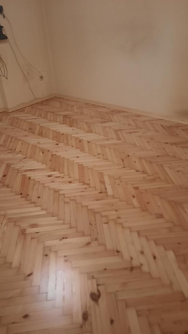 Jasny parkiet ułożony w jodełkę w pokoju z beżowymi ścianami. Widoczne gniazdka elektryczne przy podłodze. Świeżo położona podłoga.