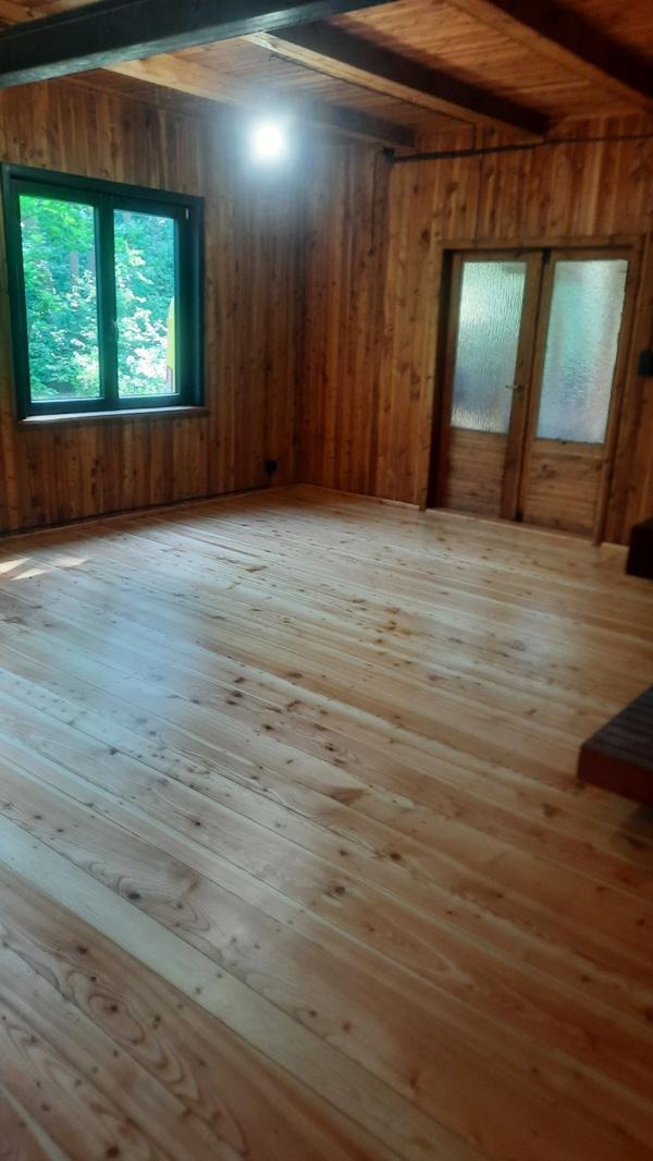 Jasne, drewniane deski podłogowe w pokoju wyłożonym boazerią; widoczne okno z zielonym widokiem i drewniane drzwi z szybami. Naturalne światło.