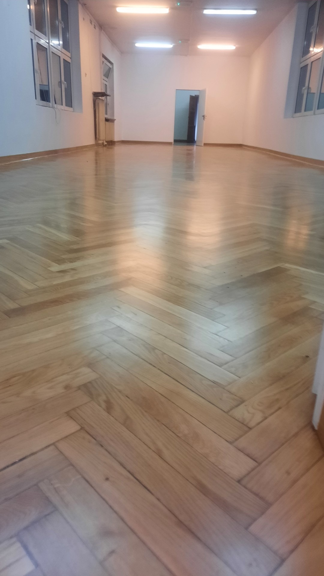 Jasny, odnowiony parkiet ułożony w jodełkę w przestronnym, oświetlonym pomieszczeniu z białymi ścianami i oknami. Widoczne ślady po cyklinowaniu.