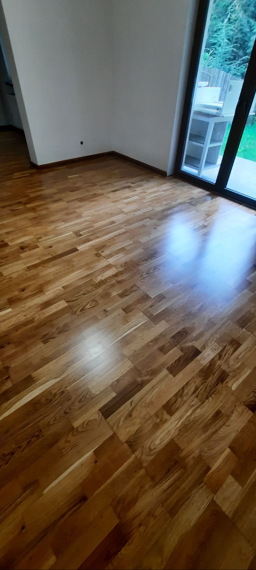 Błyszcząca, świeżo ułożona drewniana podłoga parkietowa w pustym pokoju z dużym oknem balkonowym. Widoczne naturalne usłojenie drewna.