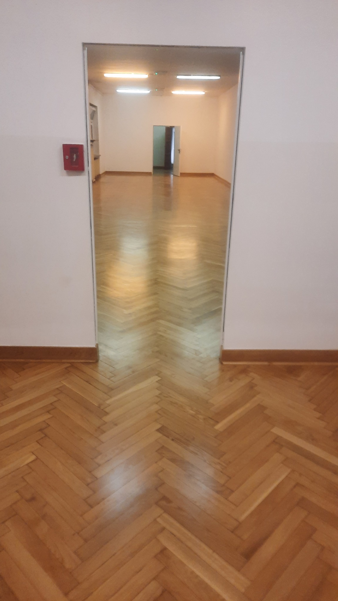 Jasny parkiet ułożony w jodełkę w pustym pomieszczeniu, widok z perspektywy drzwi. Na ścianie czerwona skrzynka alarmowa.