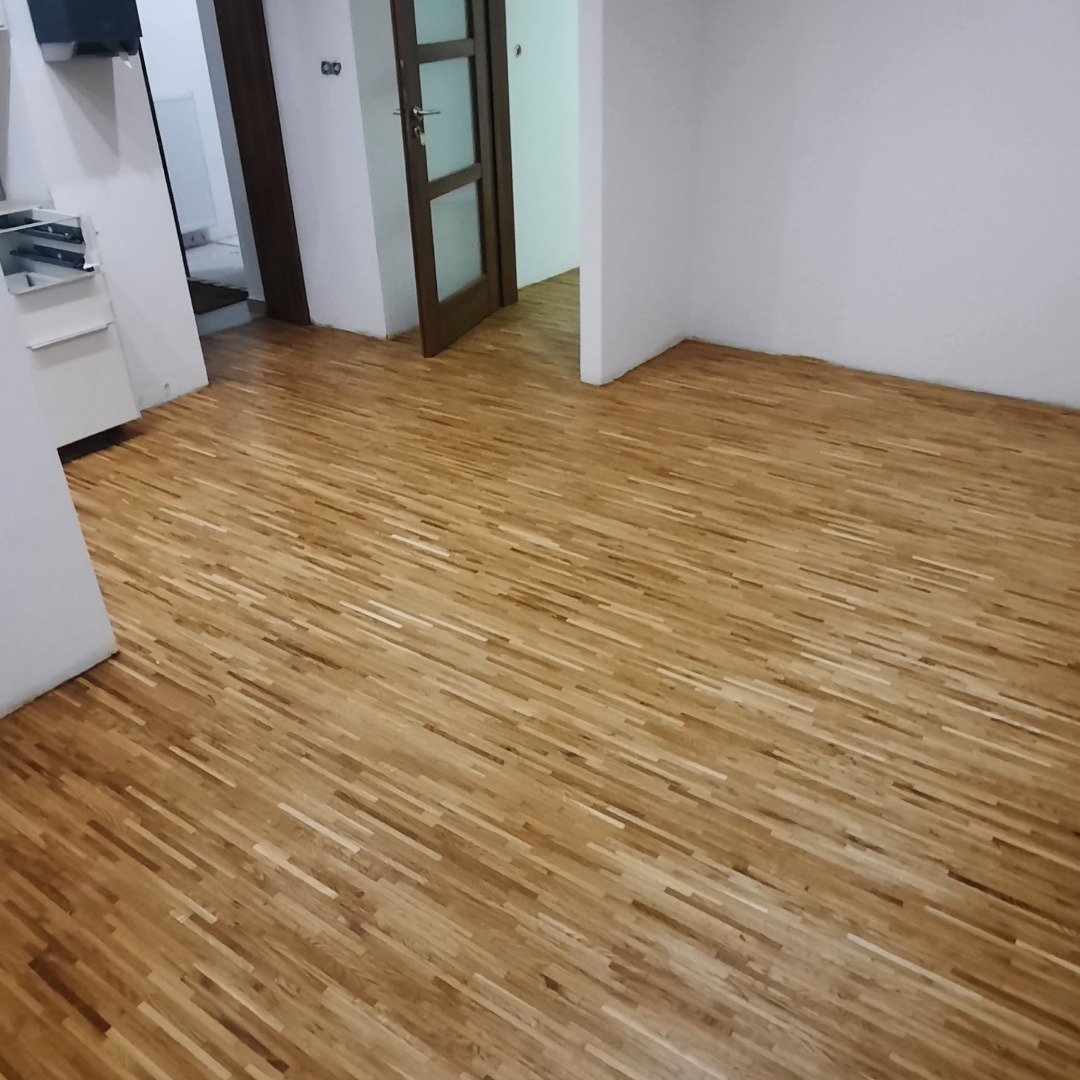 Jasny parkiet ułożony w jodełkę w pustym pokoju z białymi ścianami i brązowymi drzwiami. Widoczne przejście do kolejnego pomieszczenia.