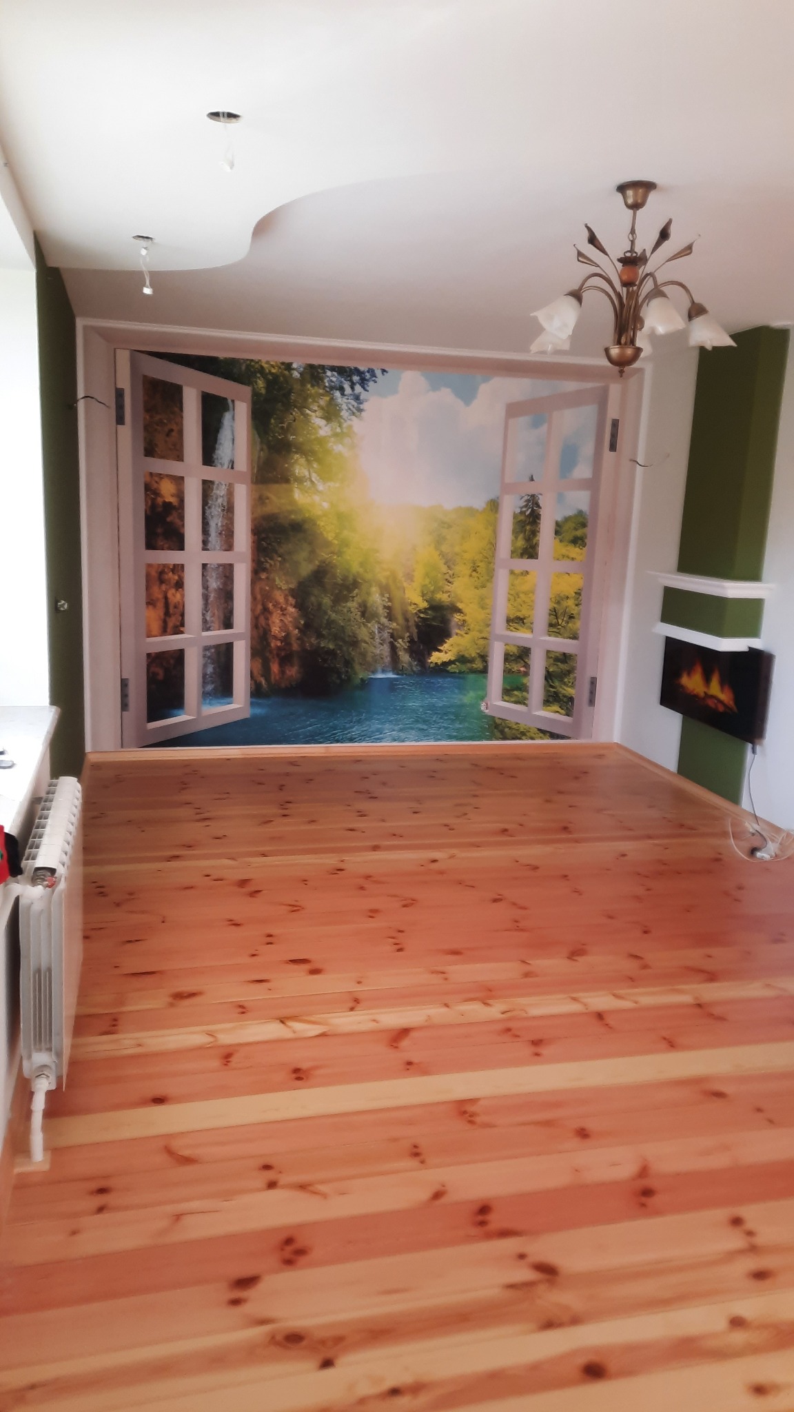 Jasne, lakierowane deski podłogowe w pustym pokoju z fototapetą okna na ścianie, ukazującą wodospad i las. Widoczny fragment grzejnika i ozdobna lampa.