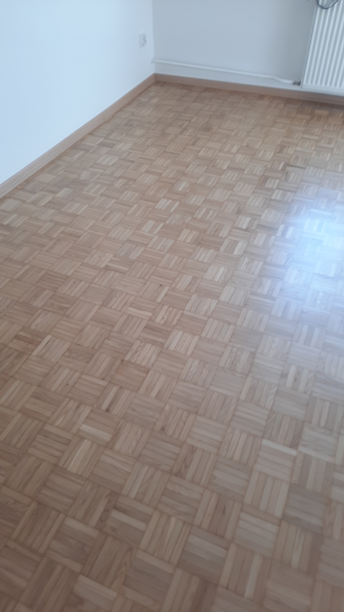 Jasny parkiet ułożony w kwadratowy wzór, widoczny fragment grzejnika i listwa przypodłogowa, ściana w tle. Perspektywa z niskiego kąta podkreśla regularność wzoru.