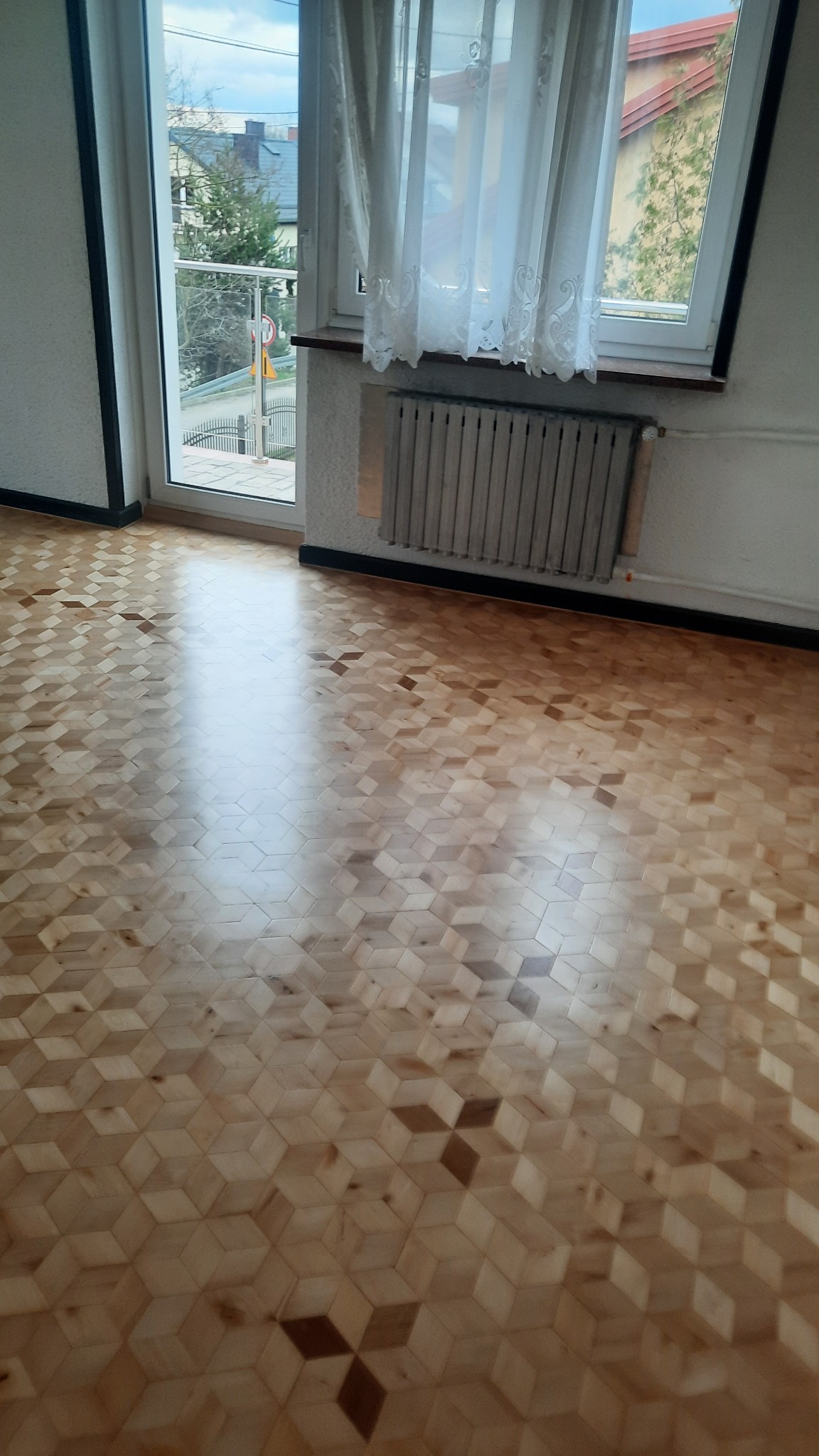 Jasny parkiet ułożony we wzór 3D z ciemniejszymi akcentami, widok z góry, okno z firanką i grzejnik w tle, refleksy światła na podłodze.