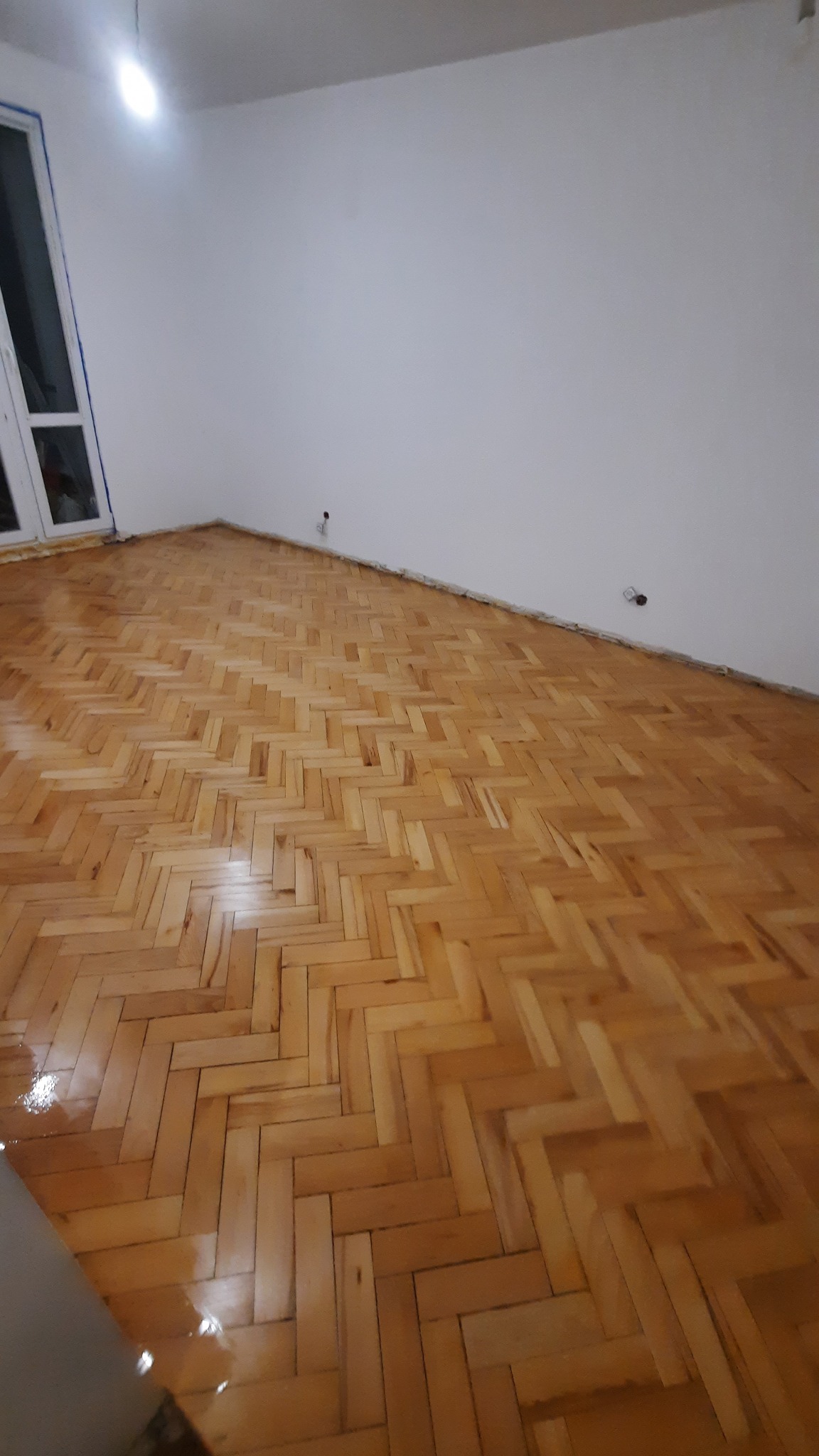 Jasny parkiet ułożony w jodełkę w pokoju z białymi ścianami. Widoczne gniazdka elektryczne. Podłoga po renowacji, błyszcząca, z widocznym refleksem światła.