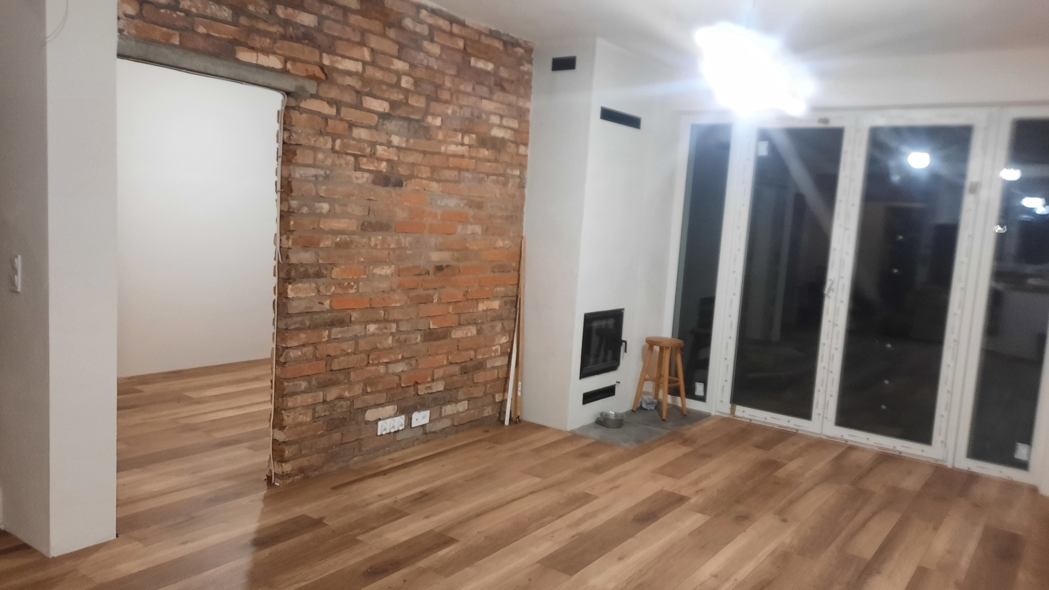 Jasne panele podłogowe w pokoju z ceglaną ścianą, białym kominkiem i dużymi oknami balkonowymi. Widoczny drewniany stołek i przejście do innego pomieszczenia.